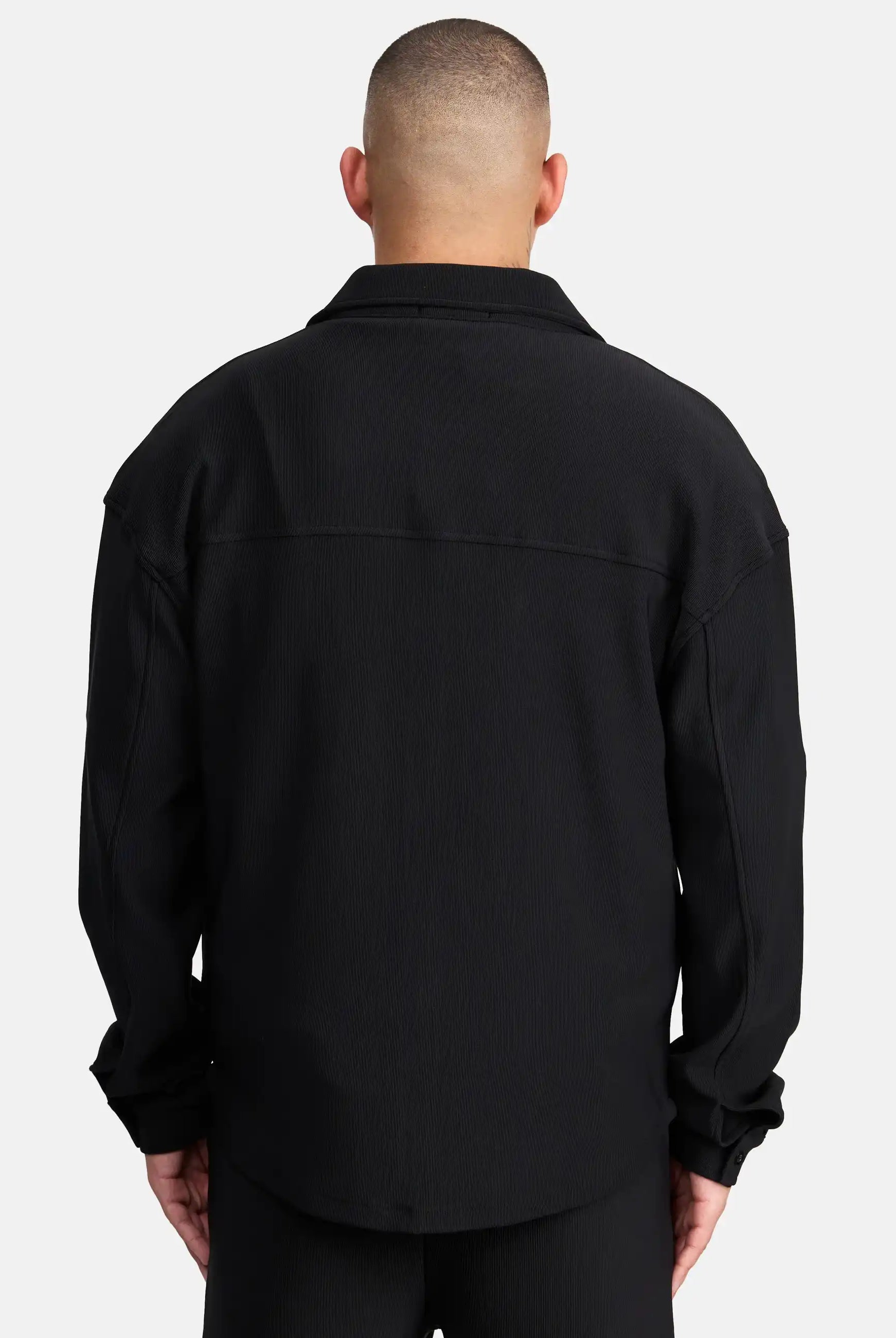 Eine Person mit kurz geschnittenem Haar wird von hinten gezeigt, die ein Trueprodigy Wyatt Oversized Langarm Hemd und eine schwarze Hose vor einem schlichten weißen Hintergrund trägt.