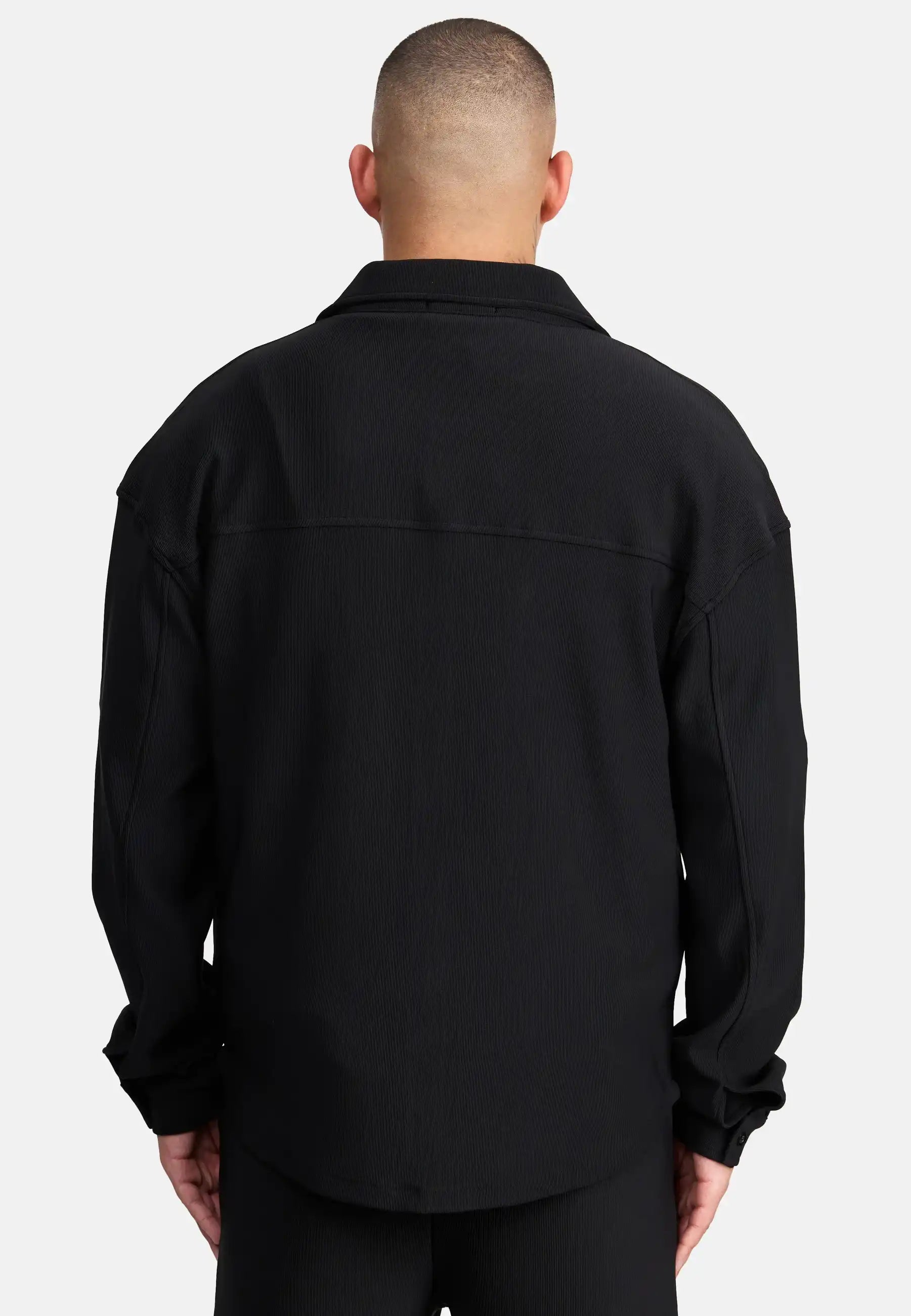 Eine Person mit kurz geschnittenem Haar wird von hinten gezeigt, die ein Trueprodigy Wyatt Oversized Langarm Hemd und eine schwarze Hose vor einem schlichten weißen Hintergrund trägt.