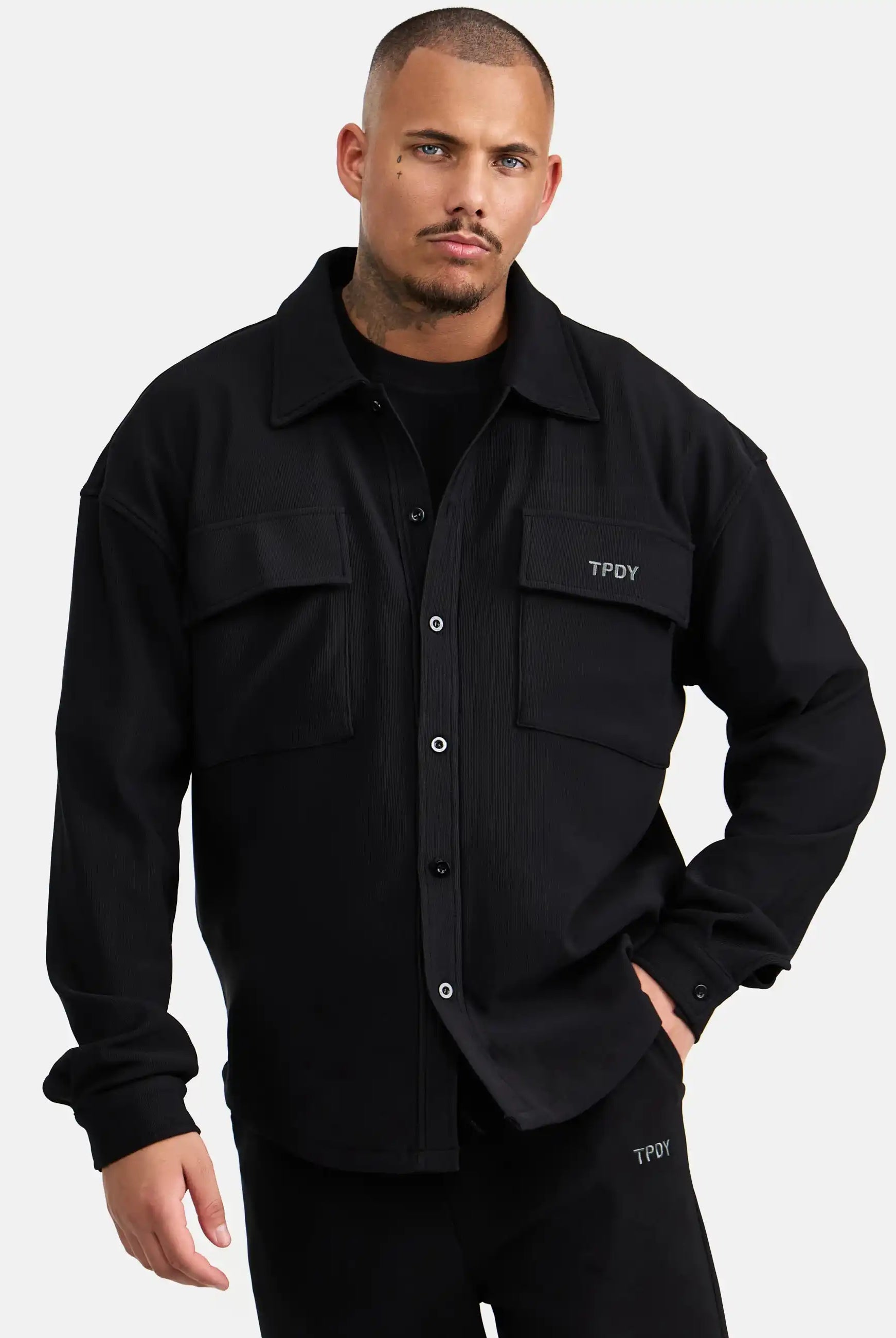 Ein Mann mit kurzen Haaren und gestutztem Bart trägt das trueprodigy Wyatt Oversized Langarm Hemd in Schwarz mit gerippter Struktur und zwei Brusttaschen, gepaart mit einer passenden schwarzen Hose, vor einem schlichten hellen Hintergrund.