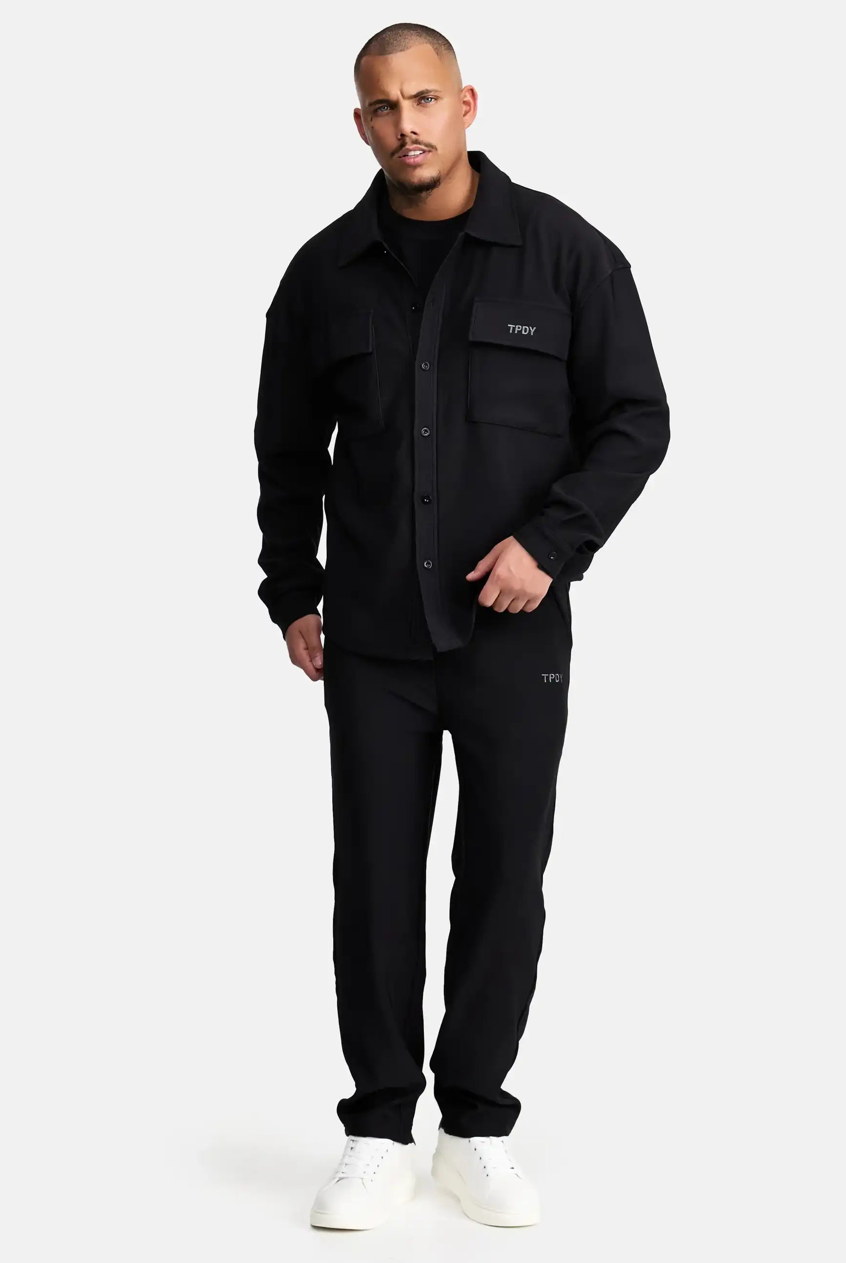 Vor einem schlichten Hintergrund trägt ein Mann mit kurzen Haaren und gepflegtem Bart das Wyatt Oversized Langarm Hemd von trueprodigy, schwarze Hose mit passenden TFOY-Logos und weiße Sneaker.