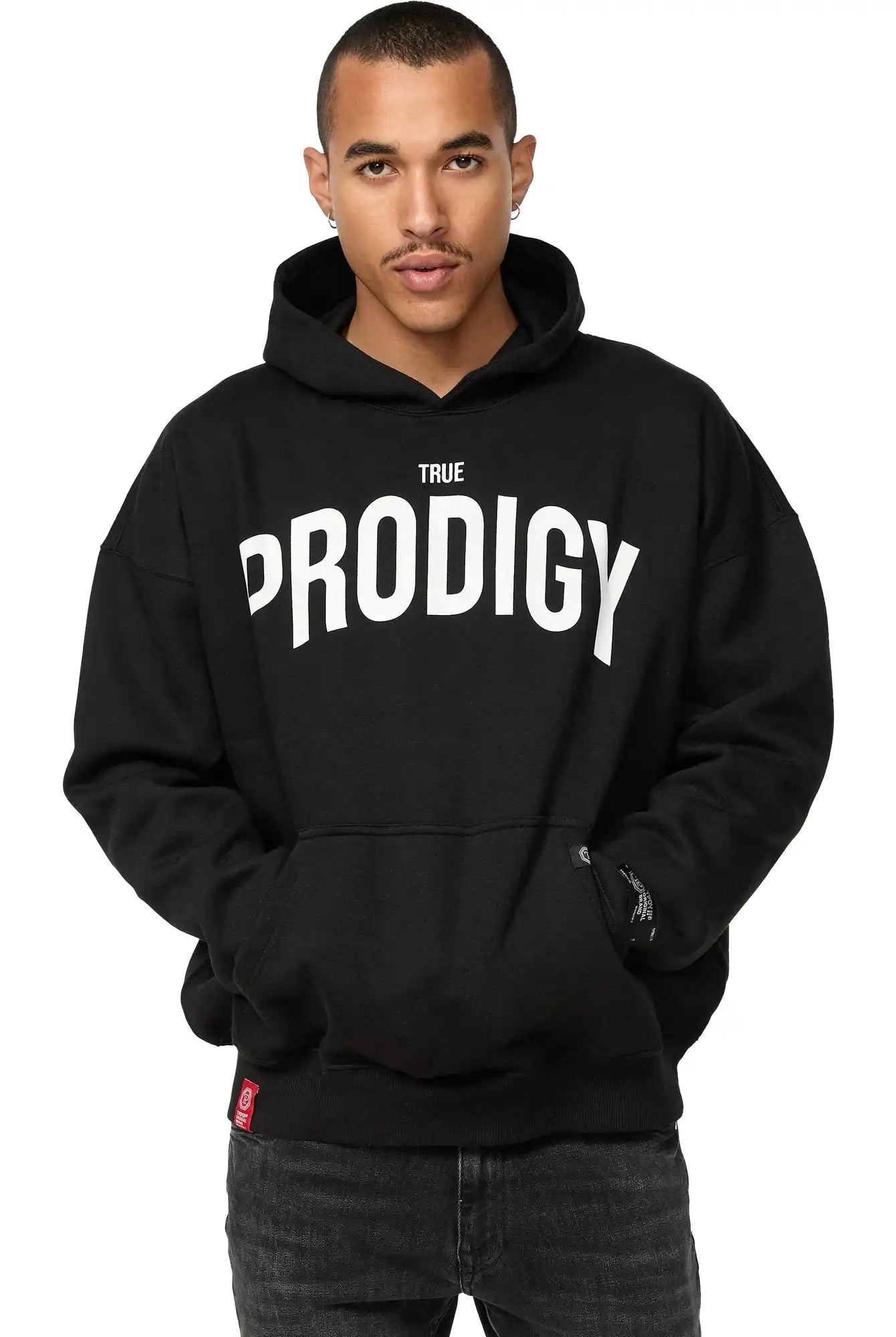 Eine Person steht mit dem Gesicht nach vorne, die Hände in den Taschen, und trägt einen schwarzen trueprodigy Tobi Oversized Hoodie mit Print mit fetten weißen TRUE PRODIGY-Schriftzügen, die den charakteristischen Oversized-Stil des Hoodies hervorheben.