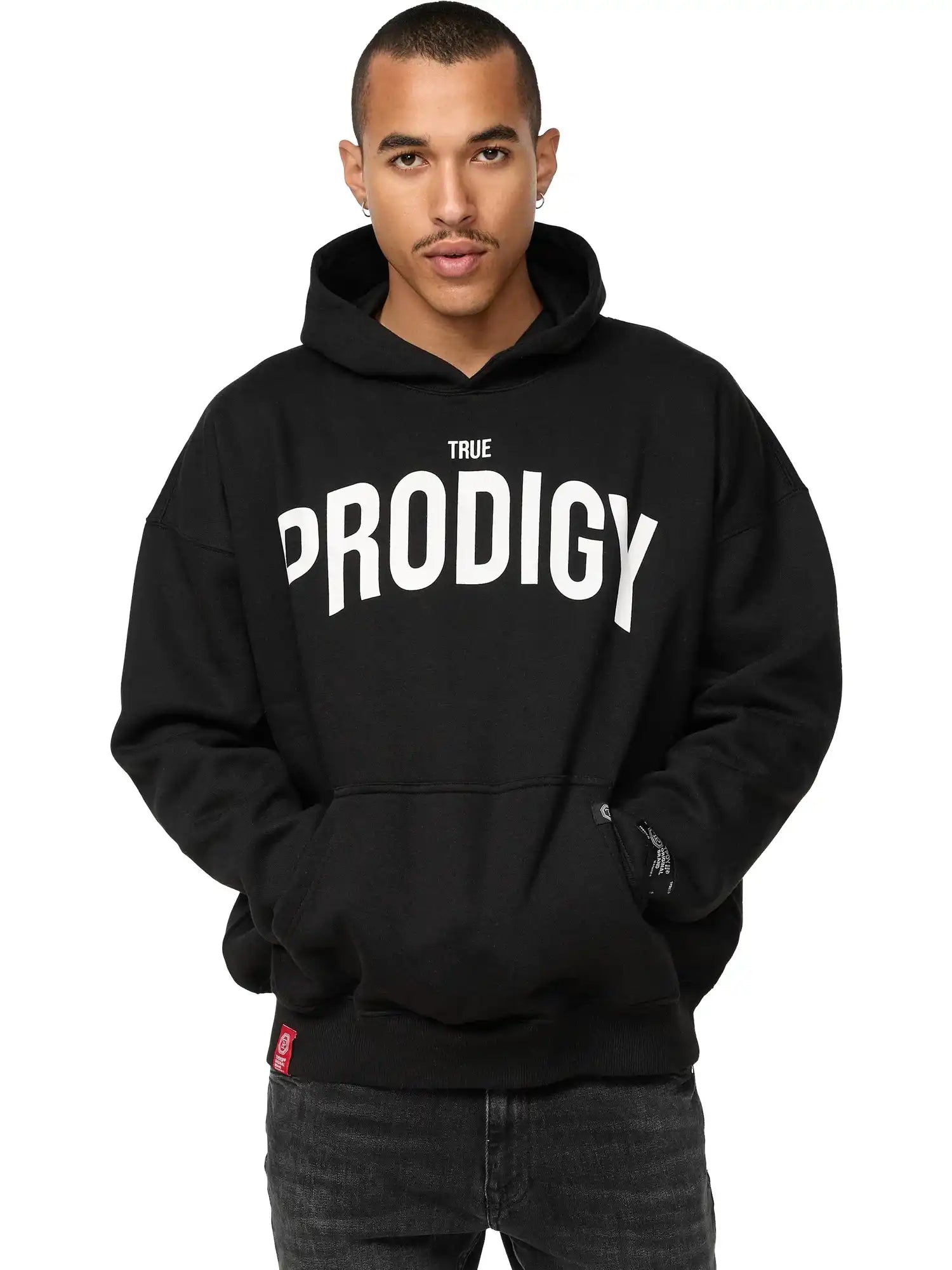 Eine Person steht mit dem Gesicht nach vorne, die Hände in den Taschen, und trägt einen schwarzen trueprodigy Tobi Oversized Hoodie mit Print mit fetten weißen TRUE PRODIGY-Schriftzügen, die den charakteristischen Oversized-Stil des Hoodies hervorheben.