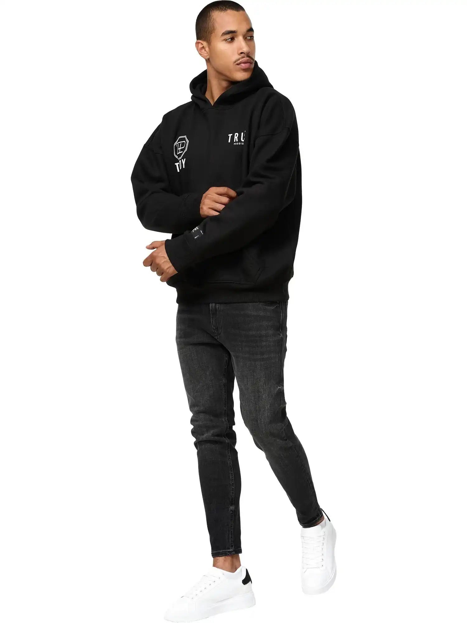 Ein Mann in einem trueprodigy Aaren Oversized Hoodie mit Print, schwarzen Skinny Jeans und weißen Sneakers steht vor einem schlichten weißen Hintergrund.
