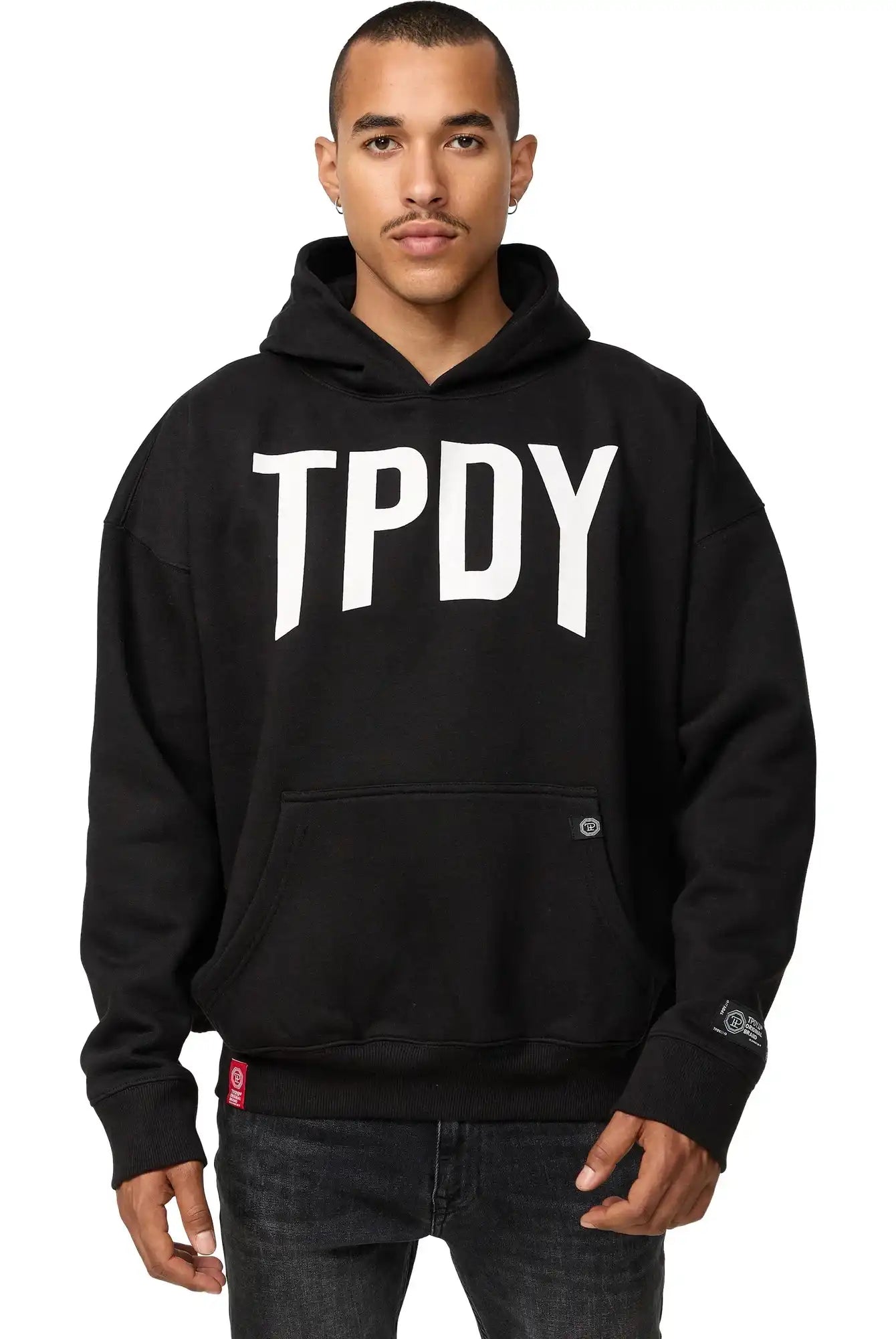 Eine Person mit kurzen Haaren und neutralem Gesichtsausdruck steht vor der Kamera, die Hände an der Kängurutasche, und trägt den trueprodigy Janne Oversized Hoodie mit Print in Schwarz mit fettem "TPDY"-Schriftzug auf der Vorderseite.