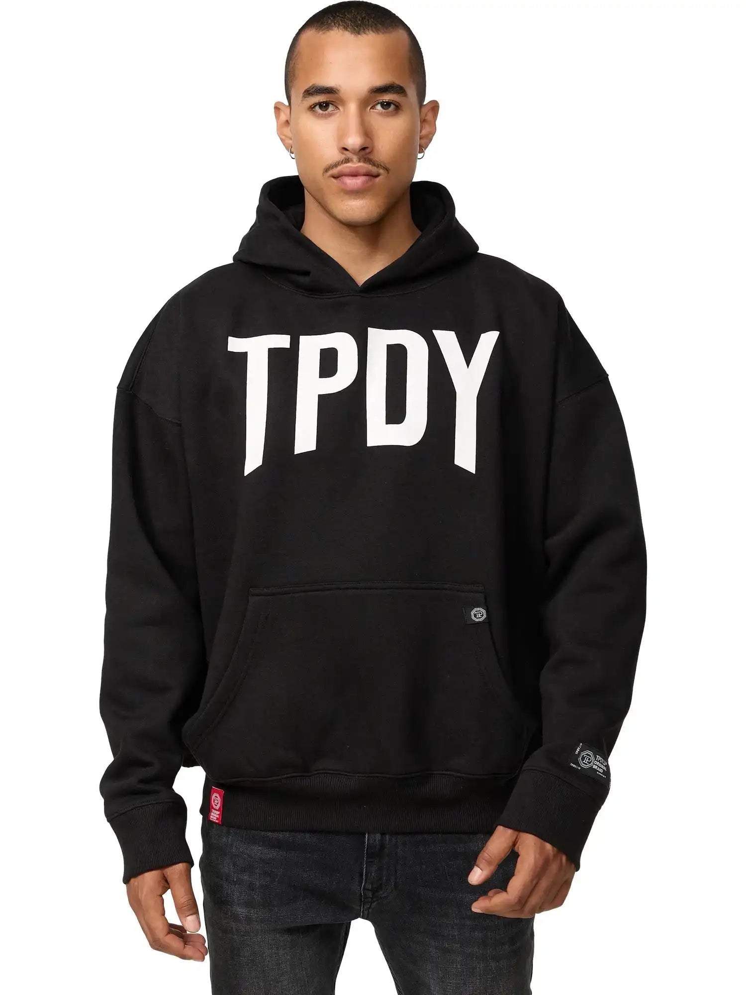 Eine Person mit kurzen Haaren und neutralem Gesichtsausdruck steht vor der Kamera, die Hände an der Kängurutasche, und trägt den trueprodigy Janne Oversized Hoodie mit Print in Schwarz mit fettem "TPDY"-Schriftzug auf der Vorderseite.