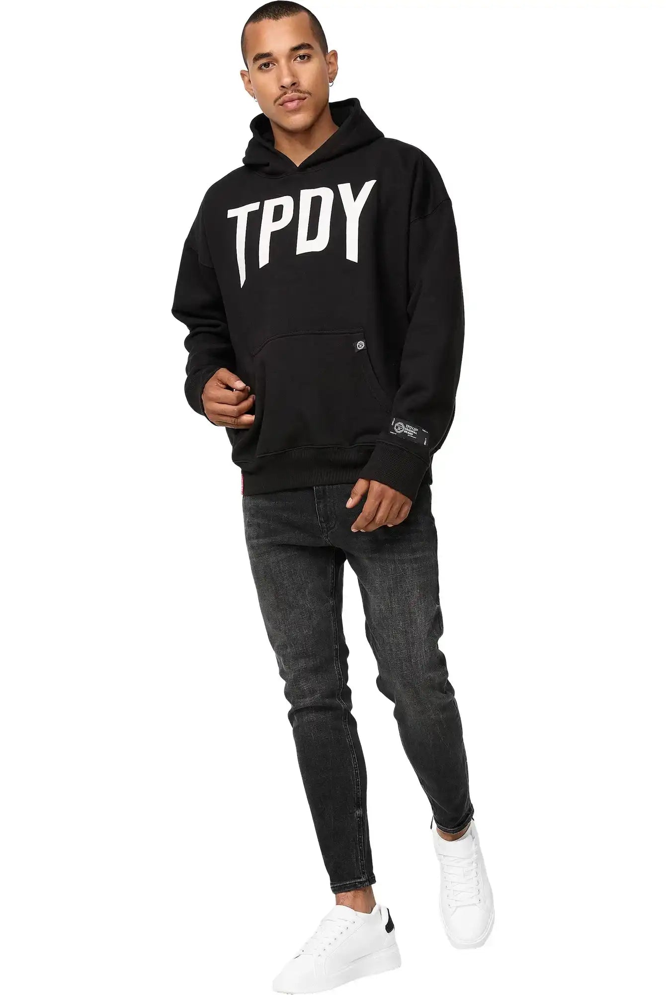 Ein Mann steht vor einem weißen Hintergrund und trägt einen trueprodigy Janne Oversized Hoodie mit Print, dunkle Skinny Jeans und weiße Turnschuhe. Er blickt selbstbewusst nach vorne und hat eine Hand in der Tasche.