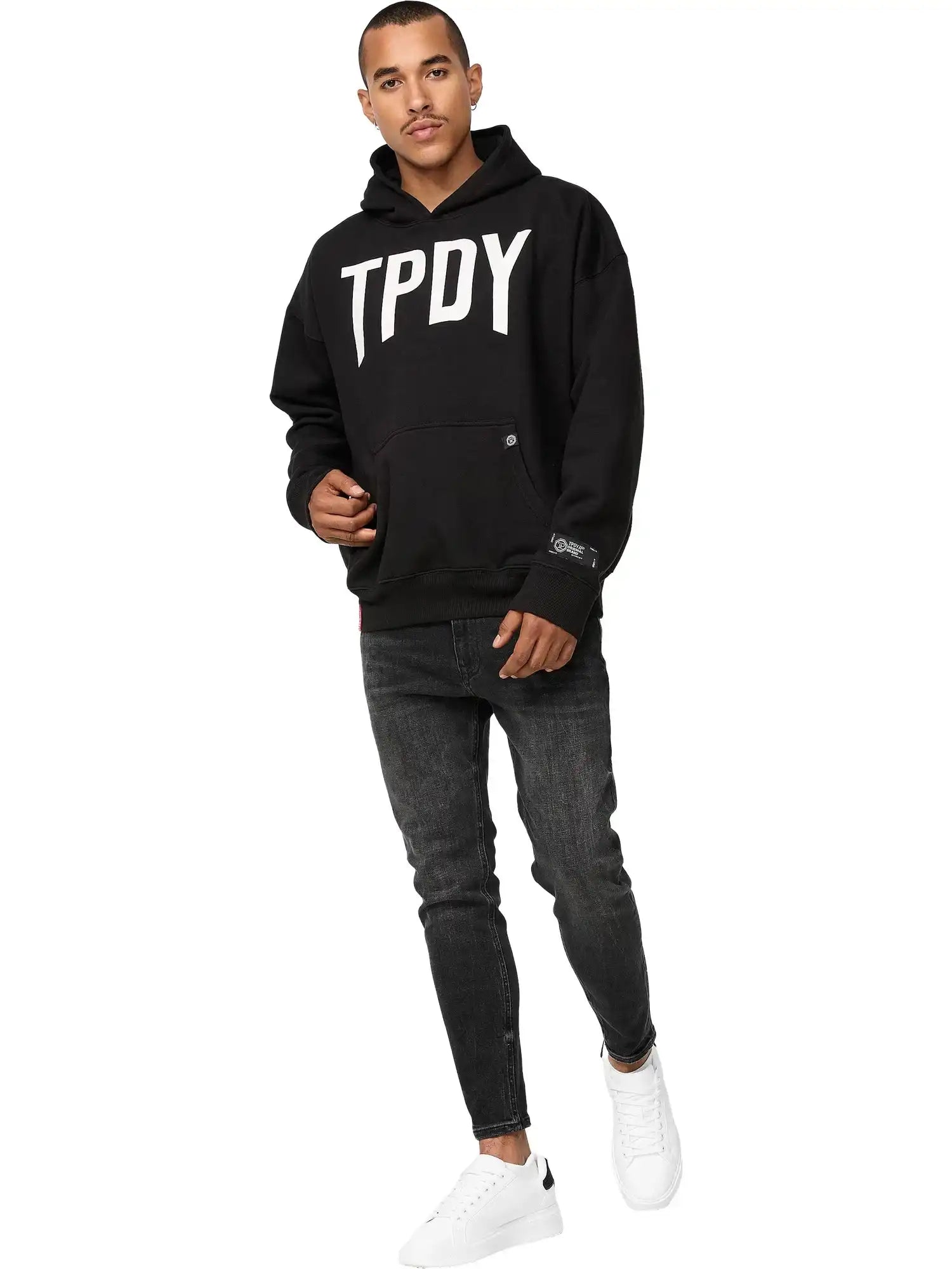 Ein Mann steht vor einem weißen Hintergrund und trägt einen trueprodigy Janne Oversized Hoodie mit Print, dunkle Skinny Jeans und weiße Turnschuhe. Er blickt selbstbewusst nach vorne und hat eine Hand in der Tasche.