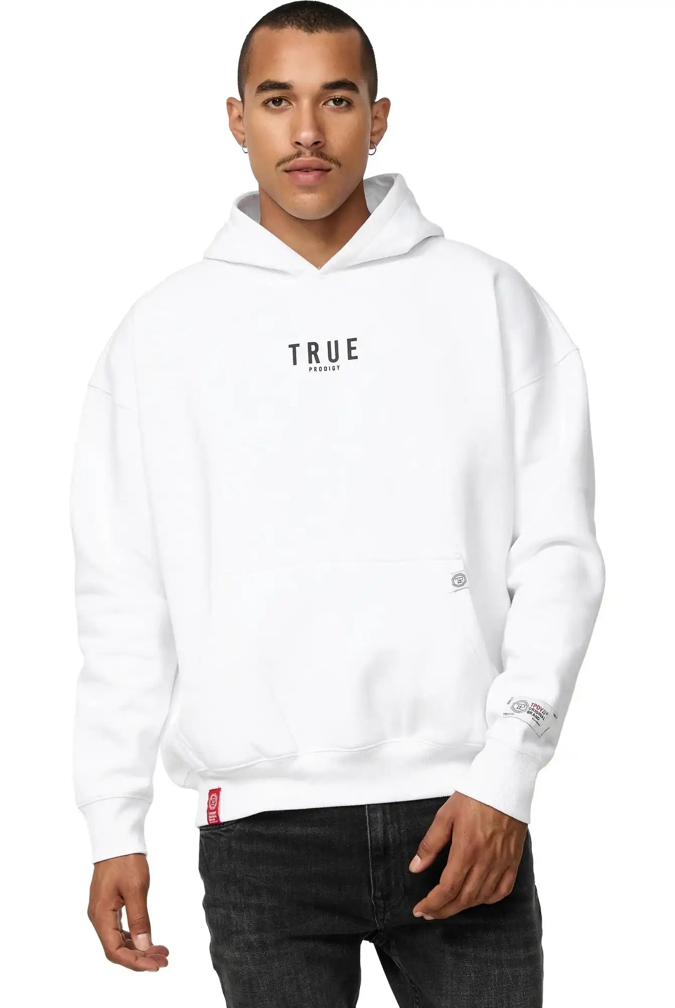 Jemand, der den Quinn Oversized Hoodie mit Print von trueprodigy mit dem Schriftzug "TRUE" auf der Vorderseite trägt, steht mit dem Gesicht nach vorne. Dieser Unisex-Kapuzenpullover hat eine moderne, lockere Passform und wird mit einer schwarzen Hose kombiniert. Die Person hat kurze Haare.