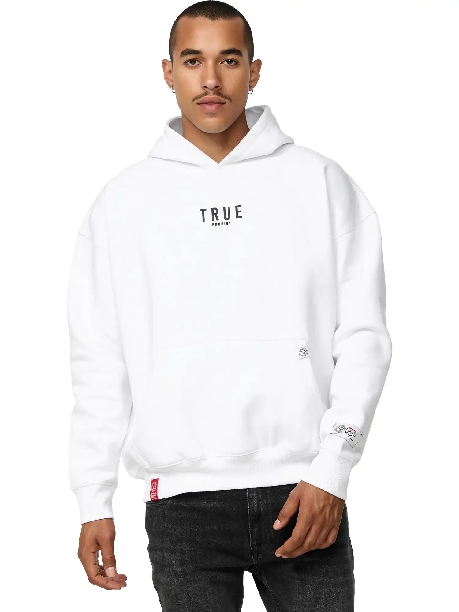 Jemand, der den Quinn Oversized Hoodie mit Print von trueprodigy mit dem Schriftzug "TRUE" auf der Vorderseite trägt, steht mit dem Gesicht nach vorne. Dieser Unisex-Kapuzenpullover hat eine moderne, lockere Passform und wird mit einer schwarzen Hose kombiniert. Die Person hat kurze Haare.
