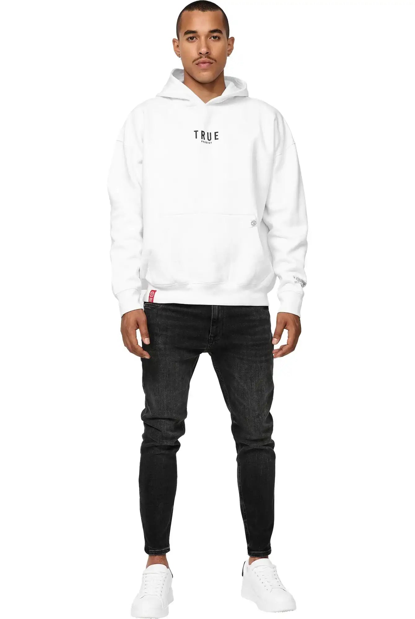 Ein Mann in einem Trueprodigy Quinn Oversized Hoodie mit Print, schwarzen Jeans und weißen Turnschuhen blickt nach vorne. Er hat kurze Haare, einen gestutzten Schnurrbart und einen neutralen Ausdruck vor einem schlichten weißen Hintergrund.