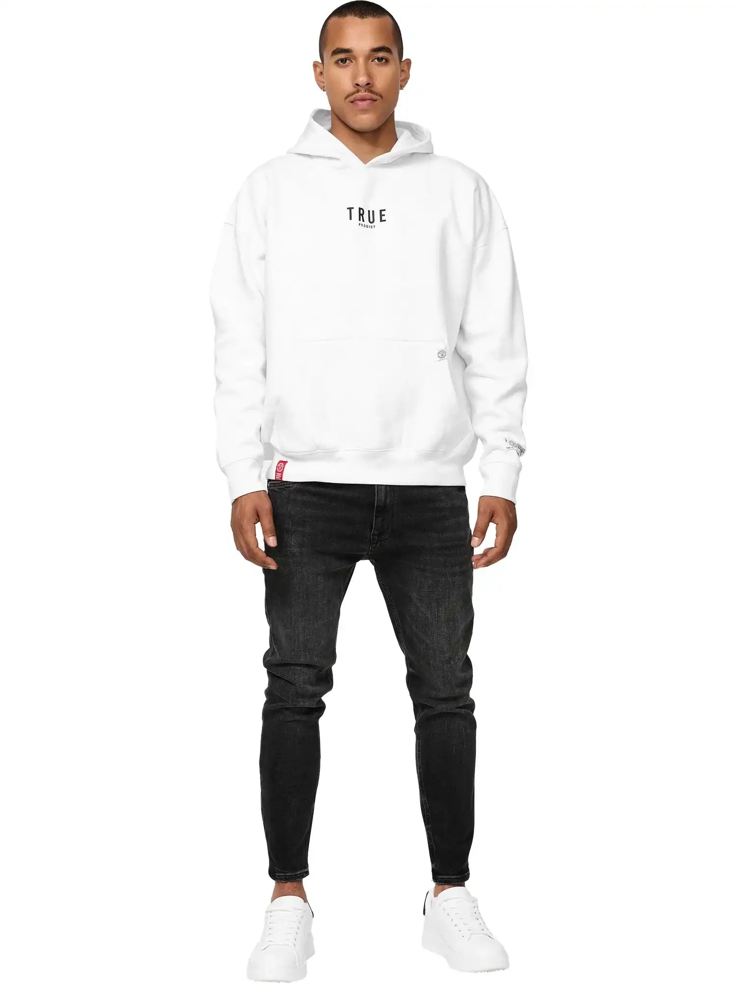 Ein Mann in einem Trueprodigy Quinn Oversized Hoodie mit Print, schwarzen Jeans und weißen Turnschuhen blickt nach vorne. Er hat kurze Haare, einen gestutzten Schnurrbart und einen neutralen Ausdruck vor einem schlichten weißen Hintergrund.