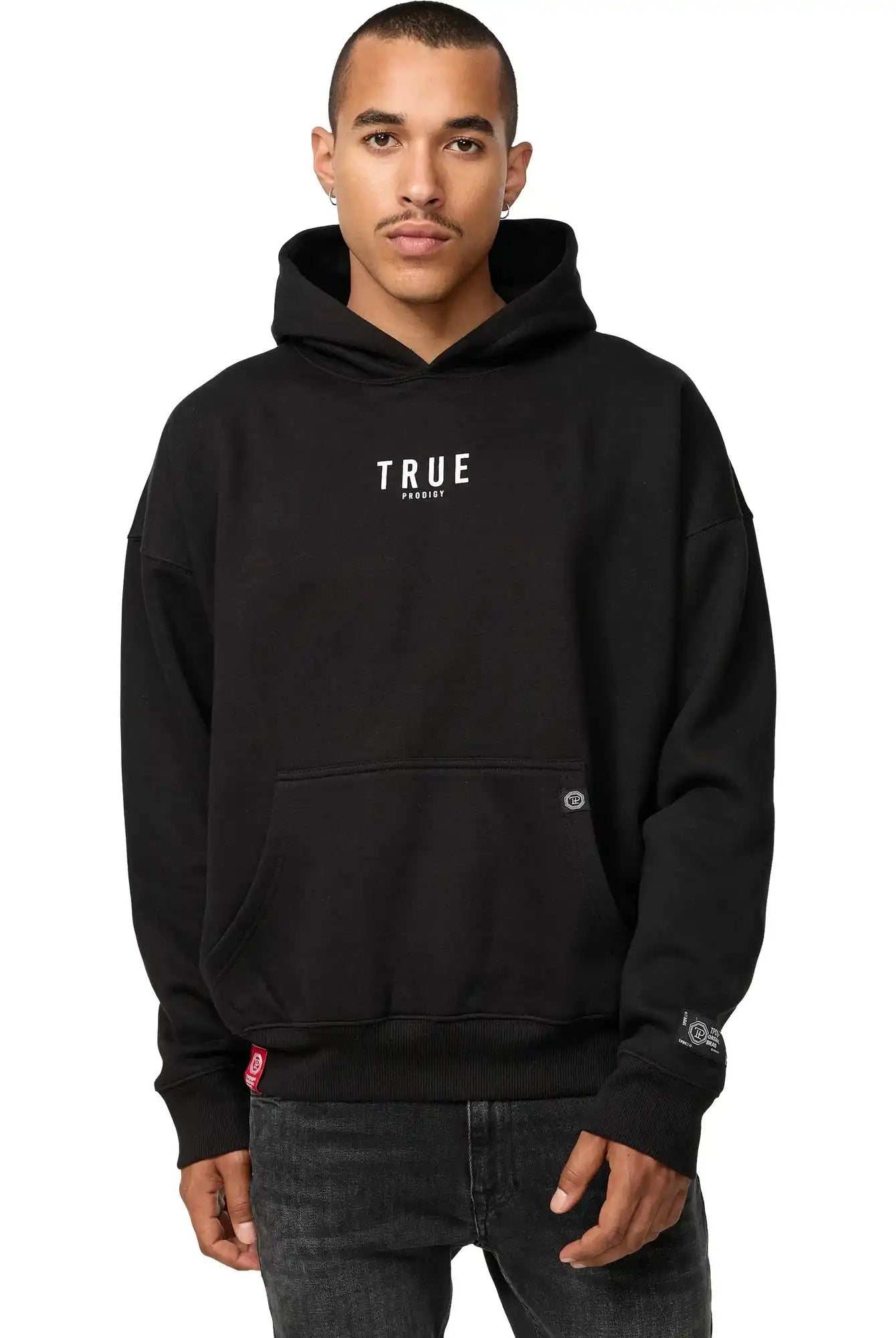 Eine Person, die einen Quinn Oversized Hoodie mit Print von trueprodigy mit dem Schriftzug "TRUE" in weißen Buchstaben auf der Vorderseite trägt, steht vor einem schlichten weißen Hintergrund und hat die Hände teilweise in der Vordertasche.