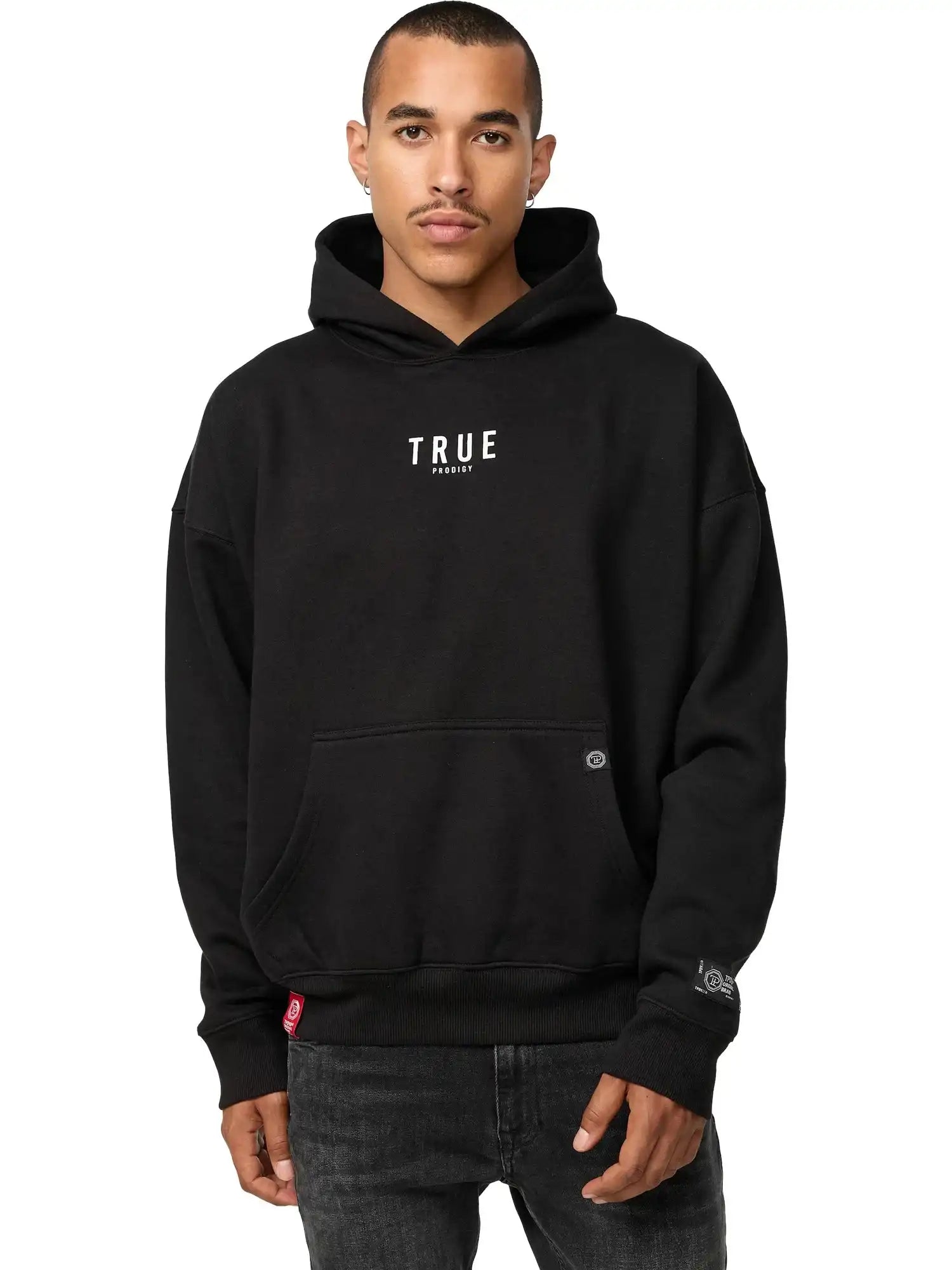 Eine Person, die einen Quinn Oversized Hoodie mit Print von trueprodigy mit dem Schriftzug "TRUE" in weißen Buchstaben auf der Vorderseite trägt, steht vor einem schlichten weißen Hintergrund und hat die Hände teilweise in der Vordertasche.