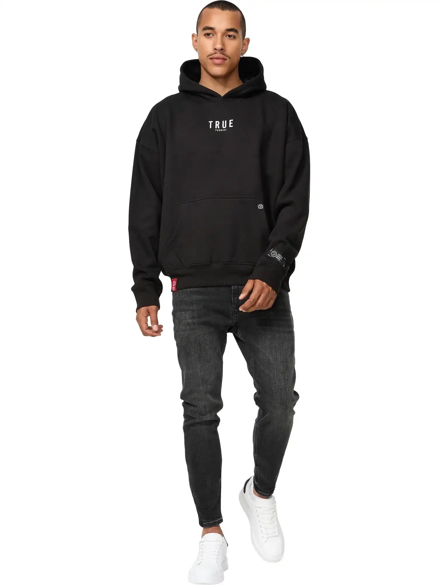 Ein Mann sieht nach vorne und trägt einen schwarzen trueprodigy Quinn Oversized Hoodie mit Print, dunkle Jeans und weiße Turnschuhe vor einem schlichten weißen Hintergrund.