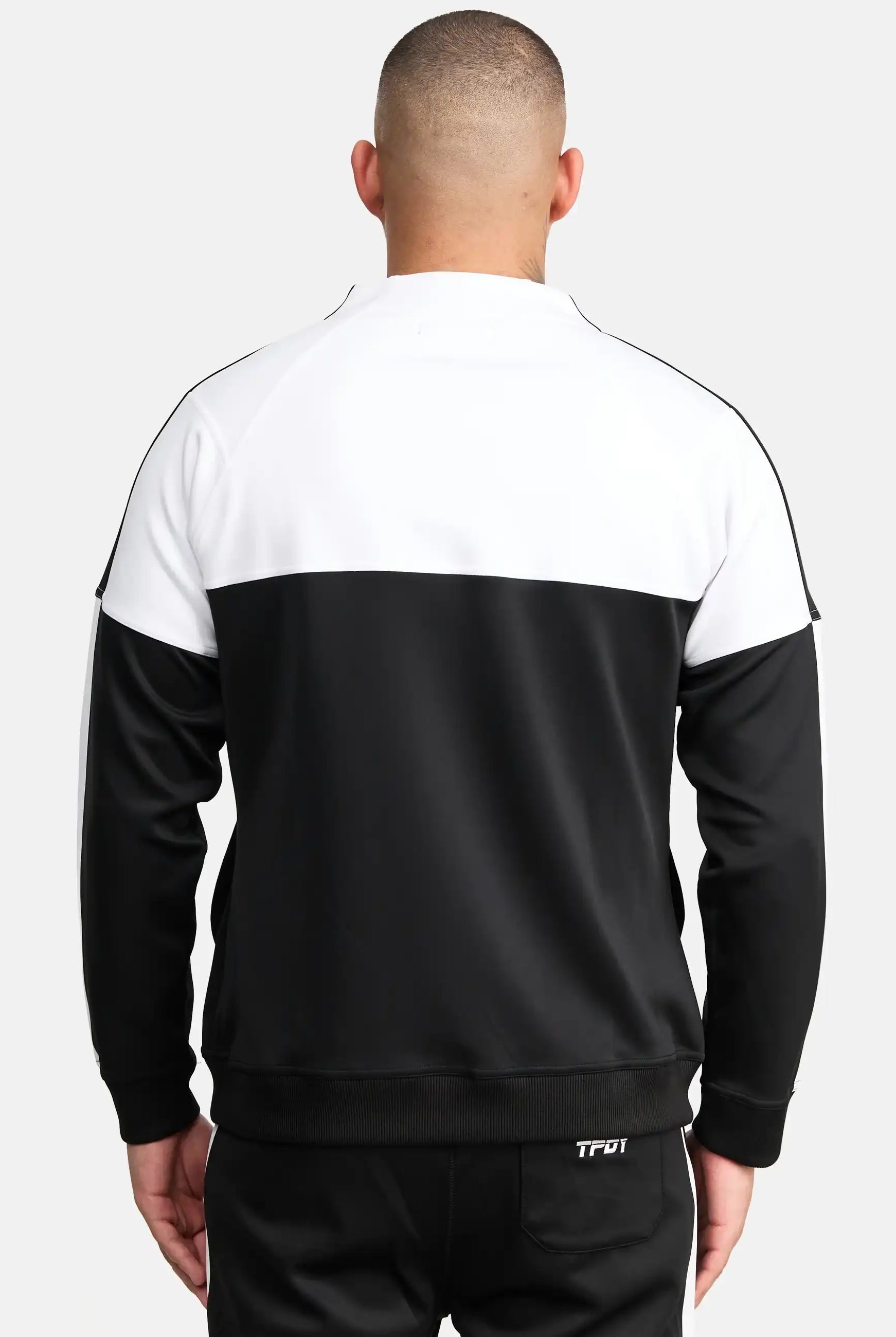 Ein Mann steht mit dem Rücken zur Kamera, trägt eine schwarze Hose und ein Trueprodigy Owen Sweatshirt mit Reißverschluss. Die Jacke hat ein weißes Oberteil, ein schwarzes Unterteil, sichtbare Nähte und Rippenbündchen.