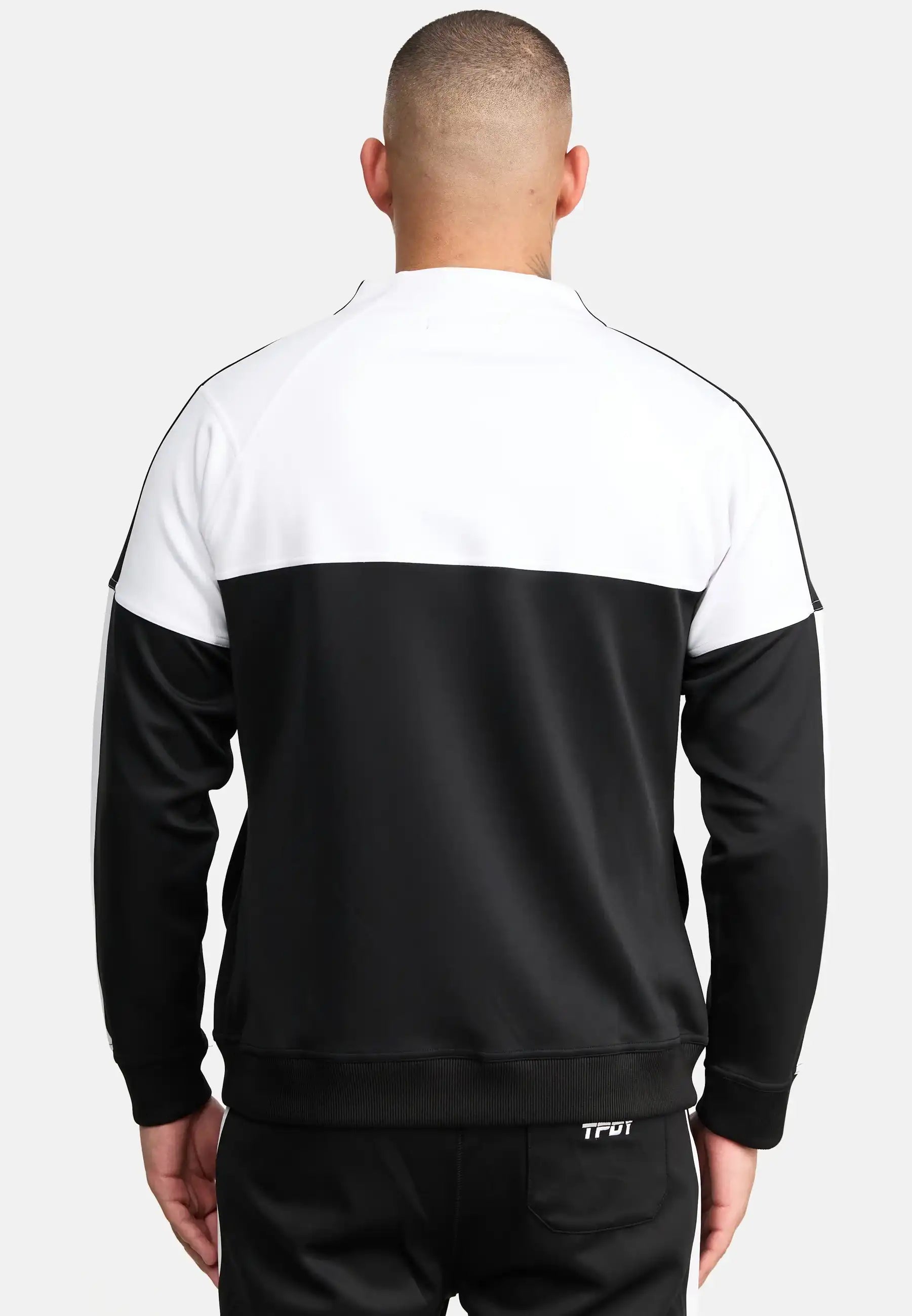 Ein Mann steht mit dem Rücken zur Kamera, trägt eine schwarze Hose und ein Trueprodigy Owen Sweatshirt mit Reißverschluss. Die Jacke hat ein weißes Oberteil, ein schwarzes Unterteil, sichtbare Nähte und Rippenbündchen.