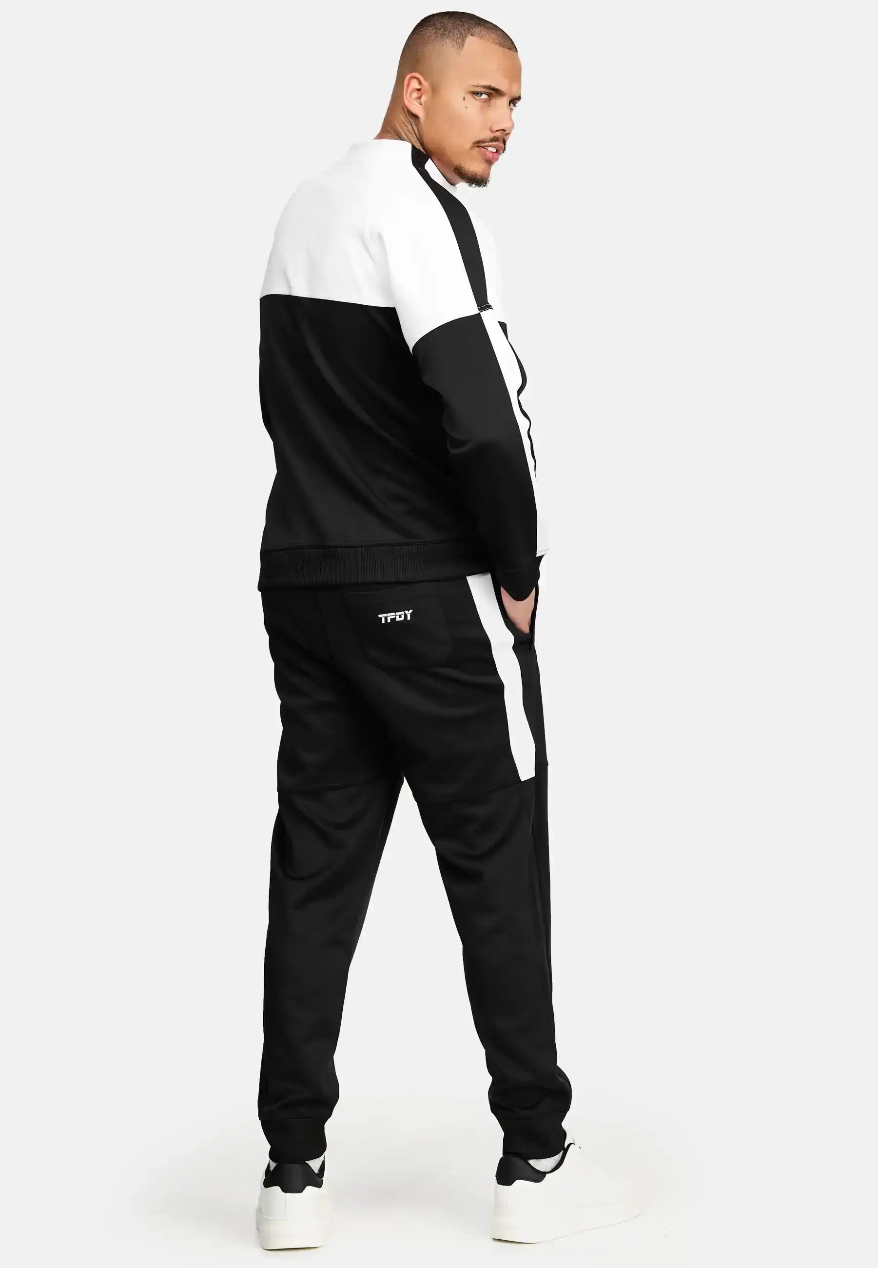 Ein Mann steht mit dem Gesicht zur Wand und schaut über seine Schulter. Er trägt einen schwarz-weißen Farbblock-Trainingsanzug mit TROY auf der Rückseite der Hose, das Owen Sweatshirt mit Reißverschluss von trueprodigy und weiße Turnschuhe vor einem schlichten Hintergrund.
