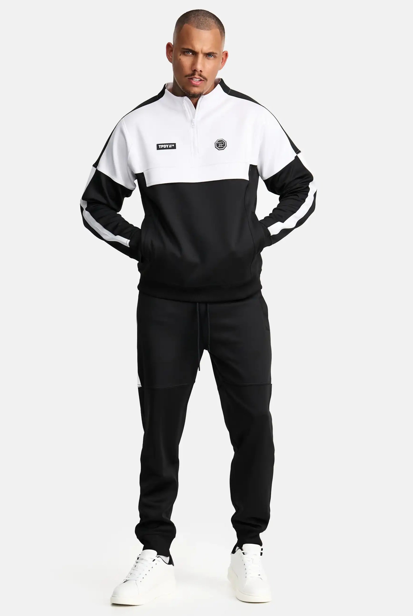 Ein Mann blickt nach vorne, hat die Hände in den Taschen und trägt das trueprodigy Owen Sweatshirt mit Reißverschluss und weiße Turnschuhe vor einem schlichten, hellen Hintergrund.