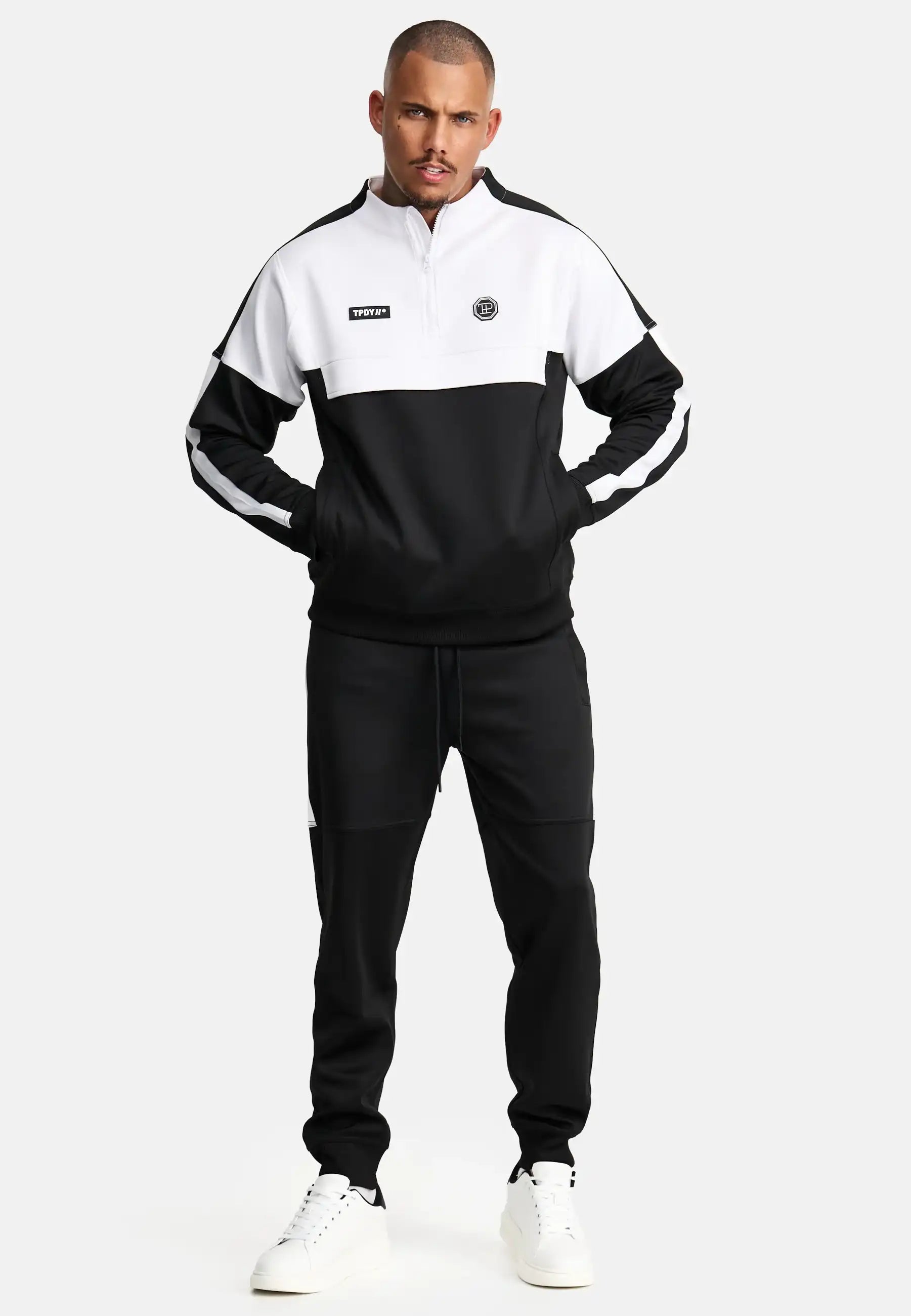 Ein Mann blickt nach vorne, hat die Hände in den Taschen und trägt das trueprodigy Owen Sweatshirt mit Reißverschluss und weiße Turnschuhe vor einem schlichten, hellen Hintergrund.