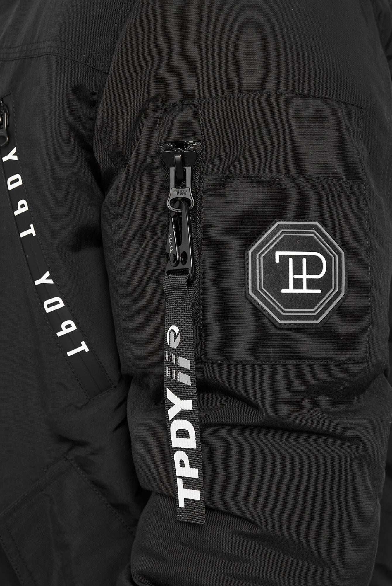 Eine Nahaufnahme des Noah Winterparka mit Kapuze und Kunstfell von trueprodigy zeigt einen schwarzen Ärmel mit einer Reißverschlusstasche, einem geometrischen Logoaufnäher und einem TPDY-Reißverschlusszug. Der weiße Text verläuft vertikal auf dem Saum, und die abnehmbare Kapuze bietet Vielseitigkeit.