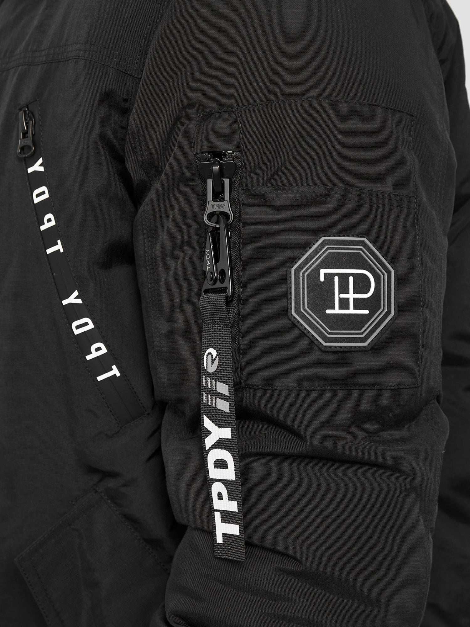 Eine Nahaufnahme des Noah Winterparka mit Kapuze und Kunstfell von trueprodigy zeigt einen schwarzen Ärmel mit einer Reißverschlusstasche, einem geometrischen Logoaufnäher und einem TPDY-Reißverschlusszug. Der weiße Text verläuft vertikal auf dem Saum, und die abnehmbare Kapuze bietet Vielseitigkeit.