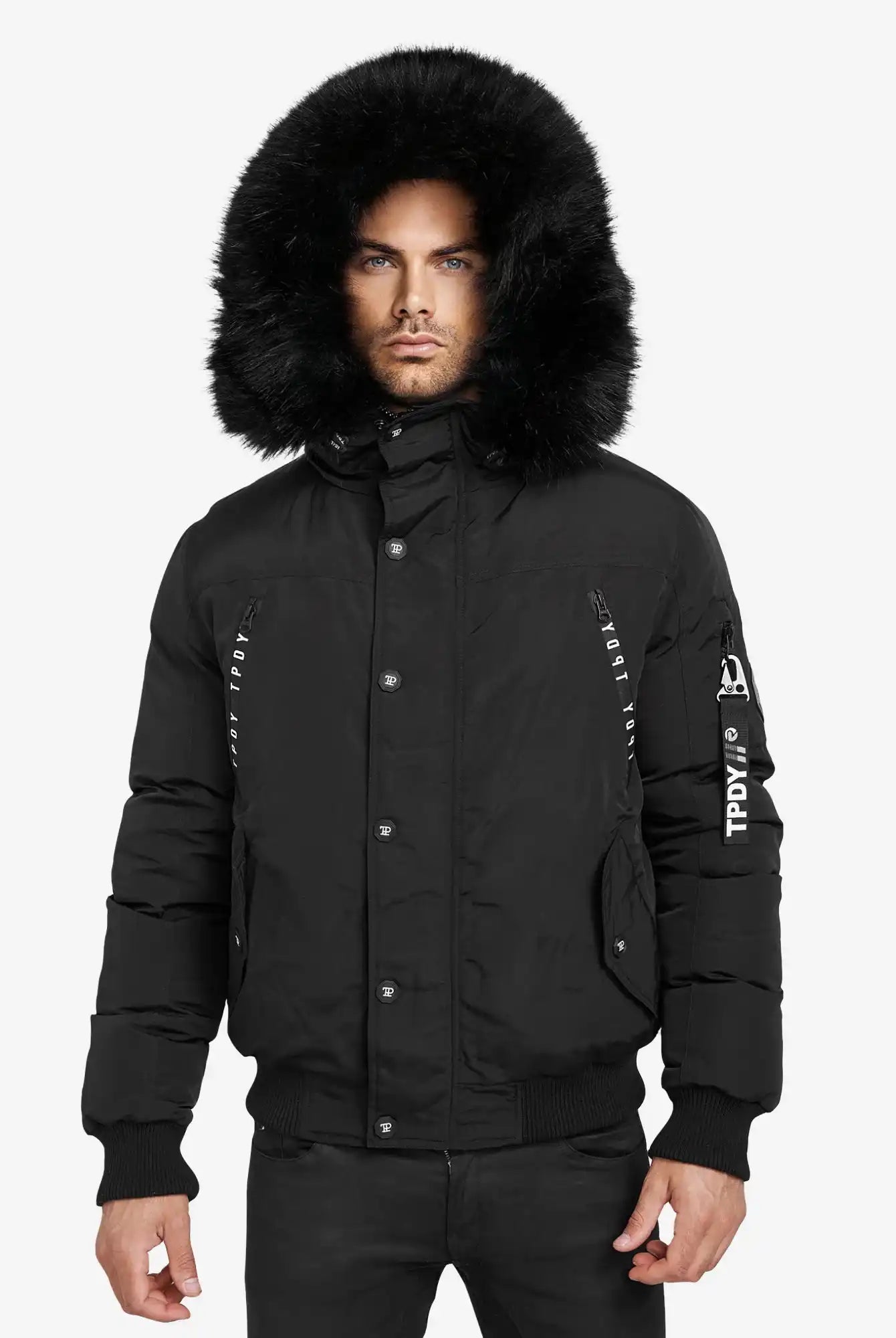 Eine Person, die den Trueprodigy Noah Winterparka mit Kapuze und Kunstfell in Schwarz mit sichtbaren Knöpfen und einem Textpatch auf dem linken Ärmel trägt, steht vor einem schlichten weißen Hintergrund.