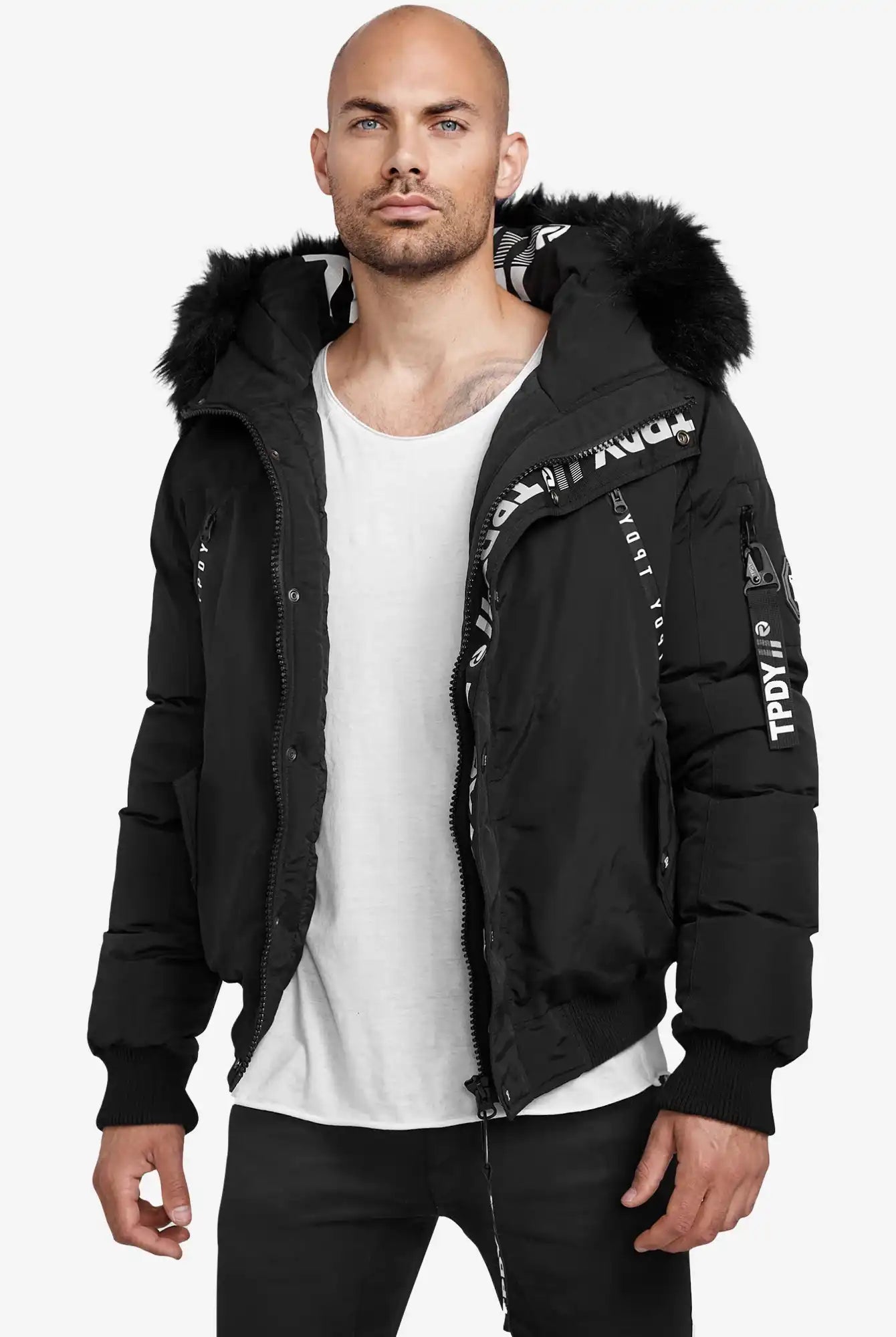 Ein Mann mit rasiertem Kopf und gestutztem Bart trägt die Trueprodigy Noah Winterparka mit Kapuze und Kunstfell, die teilweise geöffnet ist, um weiße Textdetails zu enthüllen, gepaart mit einem weißen T-Shirt und einer schwarzen Hose.