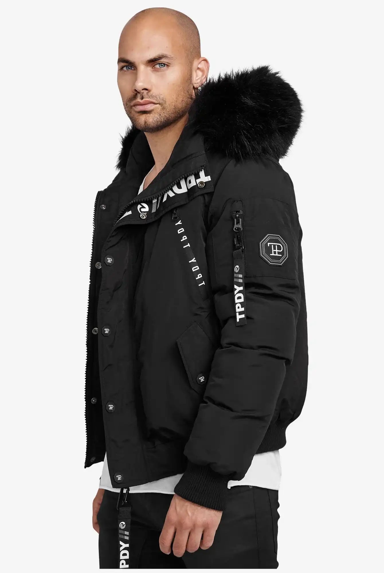 Ein Mann mit rasiertem Kopf und gestutztem Bart trägt die trueprodigy Noah Winterparka mit Kapuze und Kunstfell mit weißen Logo-Details, stehend vor einem schlichten weißen Hintergrund.