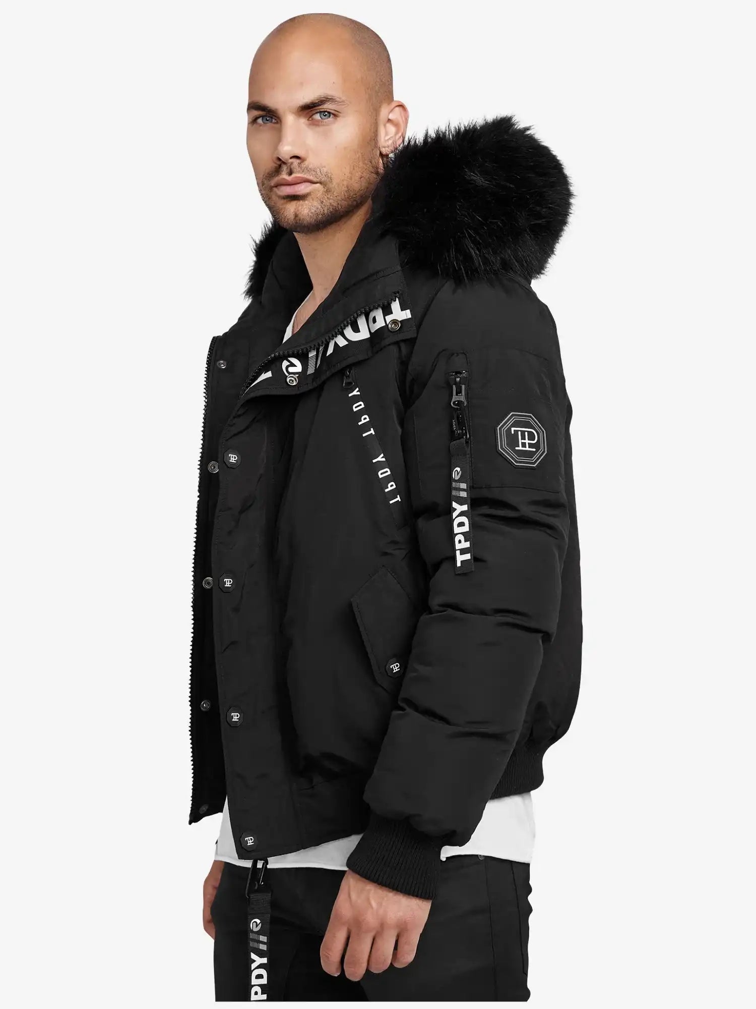 Ein Mann mit rasiertem Kopf und gestutztem Bart trägt die trueprodigy Noah Winterparka mit Kapuze und Kunstfell mit weißen Logo-Details, stehend vor einem schlichten weißen Hintergrund.