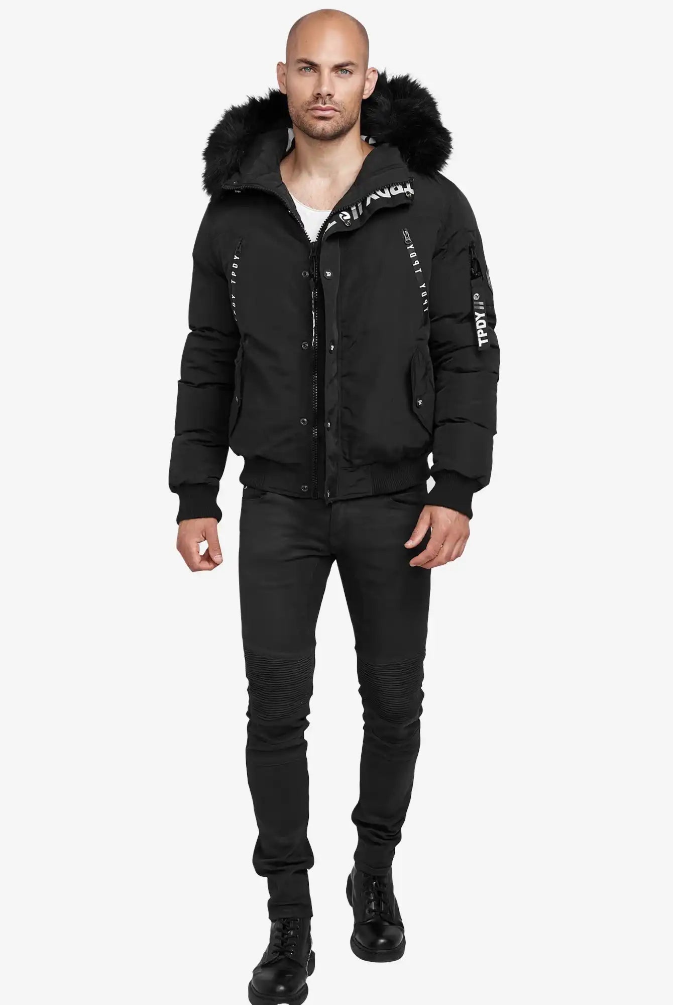 Ein Mann mit rasiertem Kopf und Bart trägt den schwarzen Noah Winterparka mit Kapuze und Kunstfell von trueprodigy, dazu schwarze Jeans und Boots. Der Parka zeigt weiße Schriftzüge auf Brust, Arm und Reißverschluss.