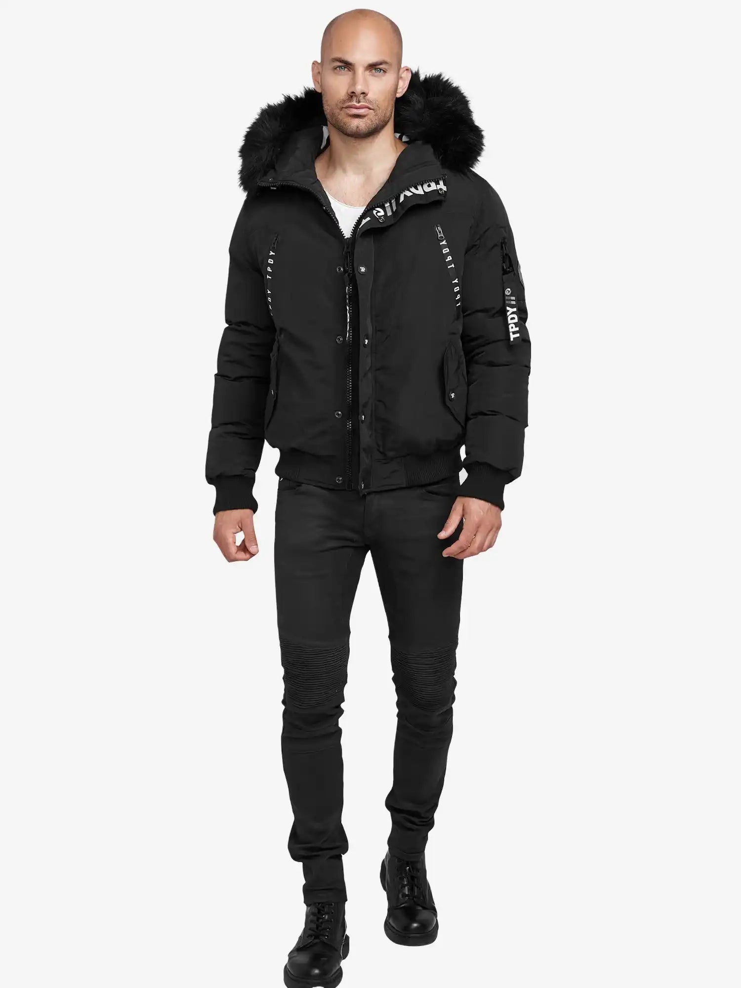 Ein Mann mit rasiertem Kopf und Bart trägt den schwarzen Noah Winterparka mit Kapuze und Kunstfell von trueprodigy, dazu schwarze Jeans und Boots. Der Parka zeigt weiße Schriftzüge auf Brust, Arm und Reißverschluss.
