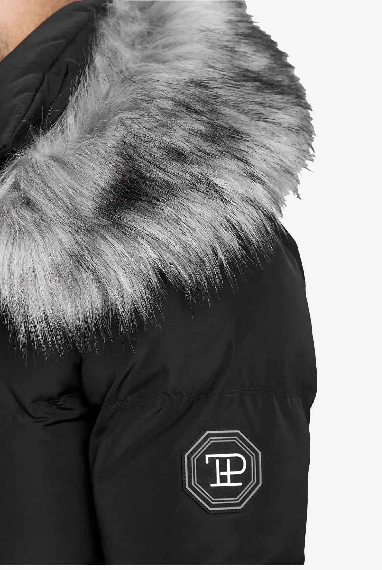 Eine Nahaufnahme der trueprodigy Zayn Winterjacke mit Kunstfell in Schwarz, mit grauer Kapuze mit Kunstfellbesatz und gesticktem, weißem, achteckigem Logopatch auf dem linken Oberarm.