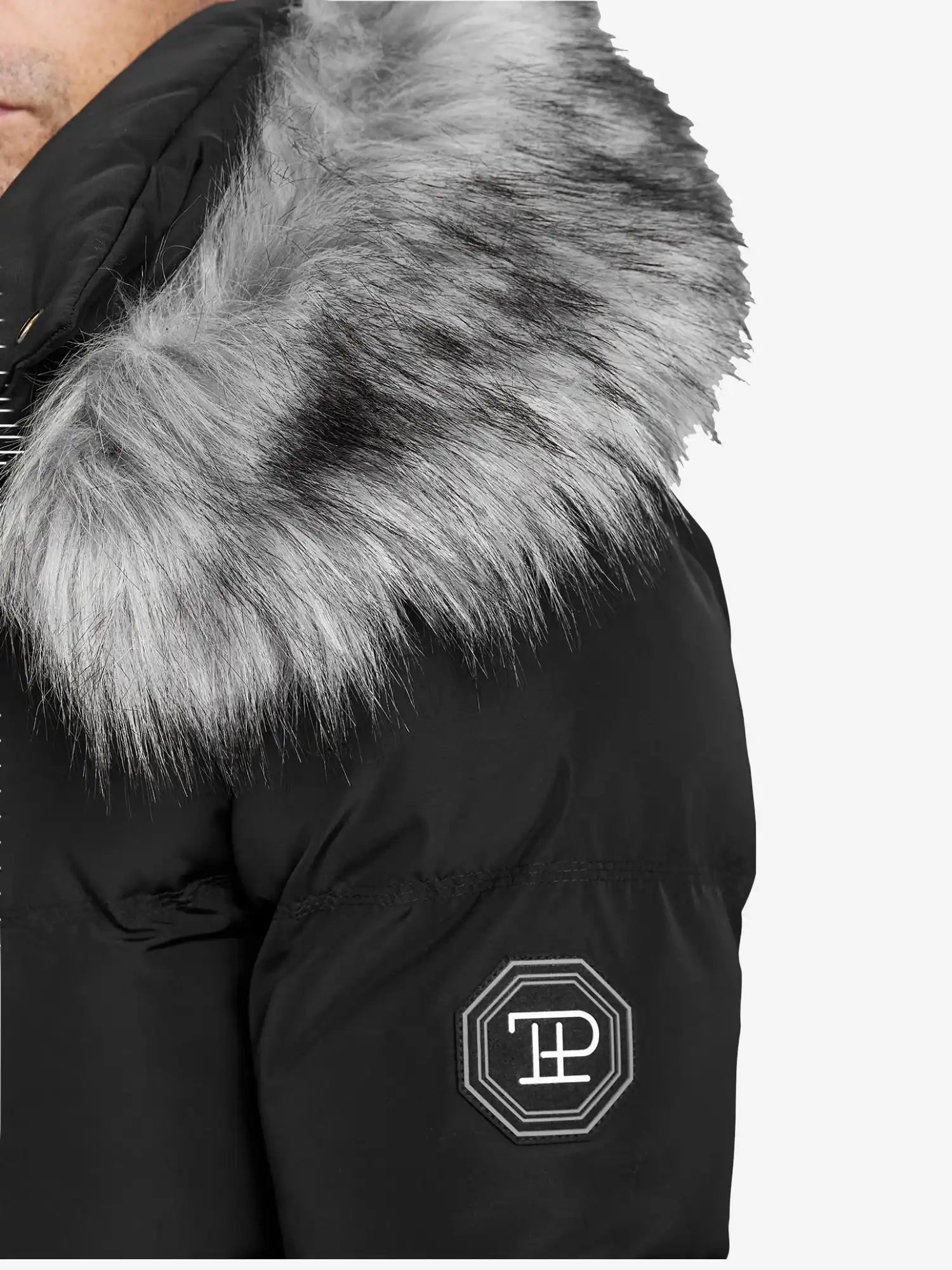Eine Nahaufnahme der trueprodigy Zayn Winterjacke mit Kunstfell in Schwarz, mit grauer Kapuze mit Kunstfellbesatz und gesticktem, weißem, achteckigem Logopatch auf dem linken Oberarm.