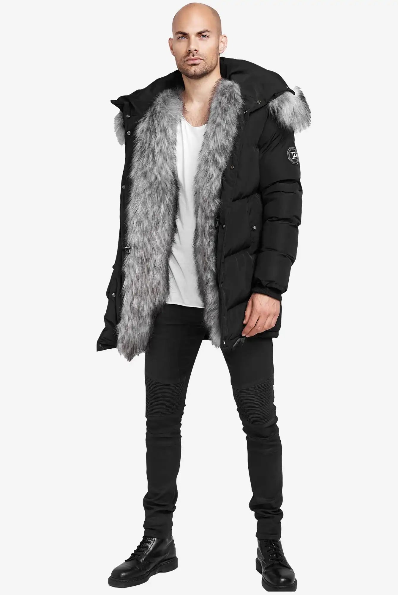 Ein Mann sieht in der schwarzen trueprodigy Zayn Winterjacke mit Kunstfell mit großem grauem Kunstfellbesatz, einem weißen T-Shirt, schwarzen Skinny Jeans und Stiefeln nach vorne. Die wasserabweisende Jacke ist mit einem Ärmelaufnäher versehen. Der Hintergrund ist einfarbig weiß.