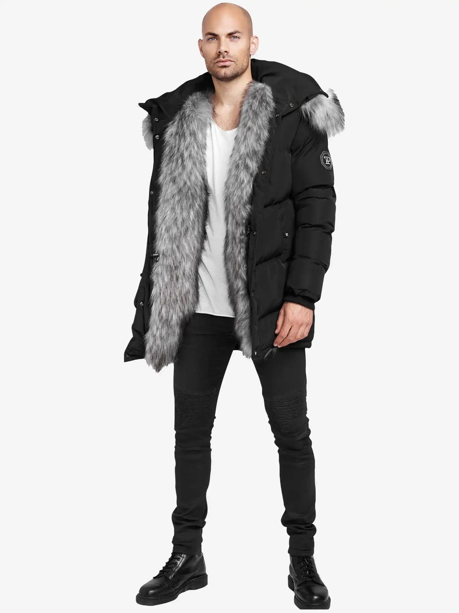 Ein Mann sieht in der schwarzen trueprodigy Zayn Winterjacke mit Kunstfell mit großem grauem Kunstfellbesatz, einem weißen T-Shirt, schwarzen Skinny Jeans und Stiefeln nach vorne. Die wasserabweisende Jacke ist mit einem Ärmelaufnäher versehen. Der Hintergrund ist einfarbig weiß.