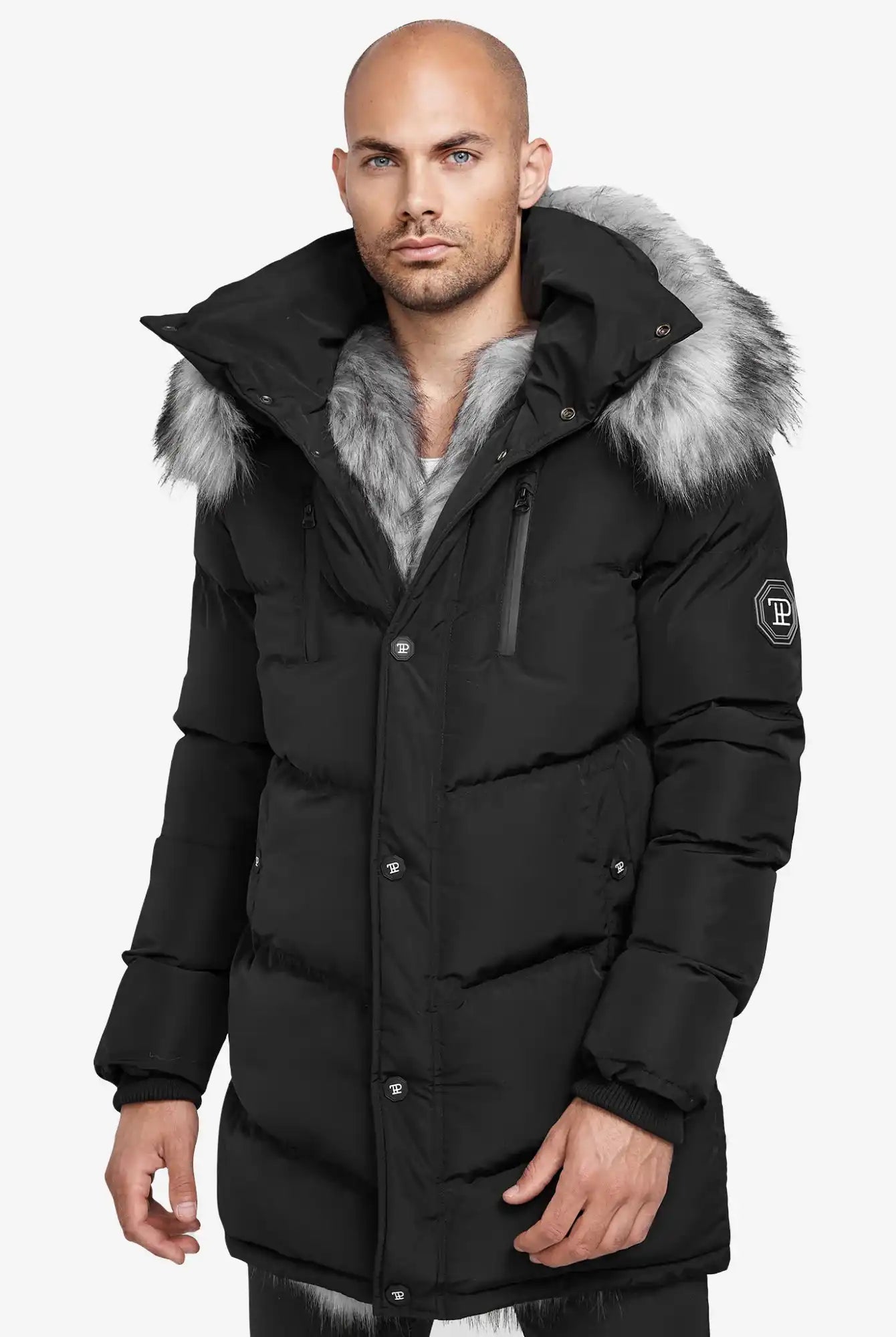 Ein Mann steht in einer schwarzen Trueprodigy Zayn Winterjacke mit Kunstfell vor der Kamera. Die Jacke hat eine große Kapuze mit Kunstpelzbesatz, sichtbare Reißverschlüsse, Knöpfe und einen Aufnäher auf dem Ärmel vor einem schlichten weißen Hintergrund.