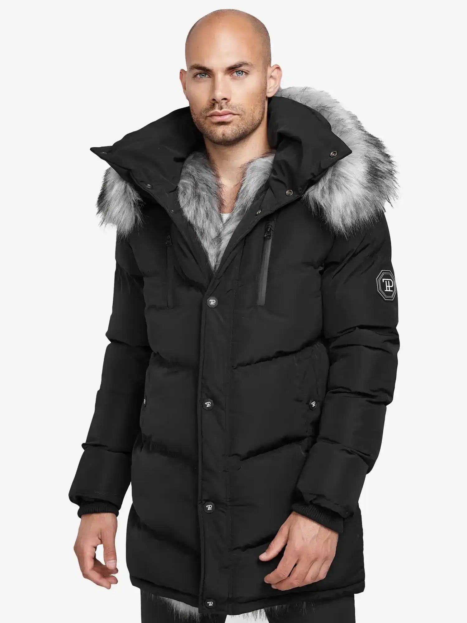 Ein Mann steht in einer schwarzen Trueprodigy Zayn Winterjacke mit Kunstfell vor der Kamera. Die Jacke hat eine große Kapuze mit Kunstpelzbesatz, sichtbare Reißverschlüsse, Knöpfe und einen Aufnäher auf dem Ärmel vor einem schlichten weißen Hintergrund.
