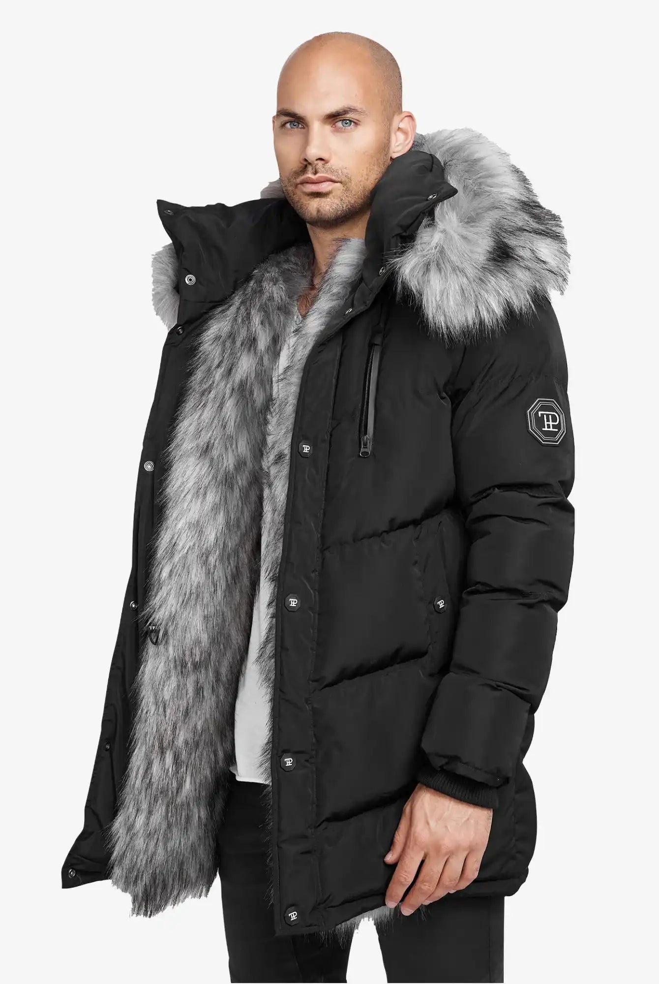 Ein Mann trägt die trueprodigy Zayn Winterjacke mit Kunstfell, einen schwarzen, wasserabweisenden Parka mit dickem grauem Kunstfell an Kapuze und Futter. Er ist mit einem Ärmelaufnäher und Druckknöpfen versehen und wird vor einem schlichten weißen Hintergrund gezeigt.