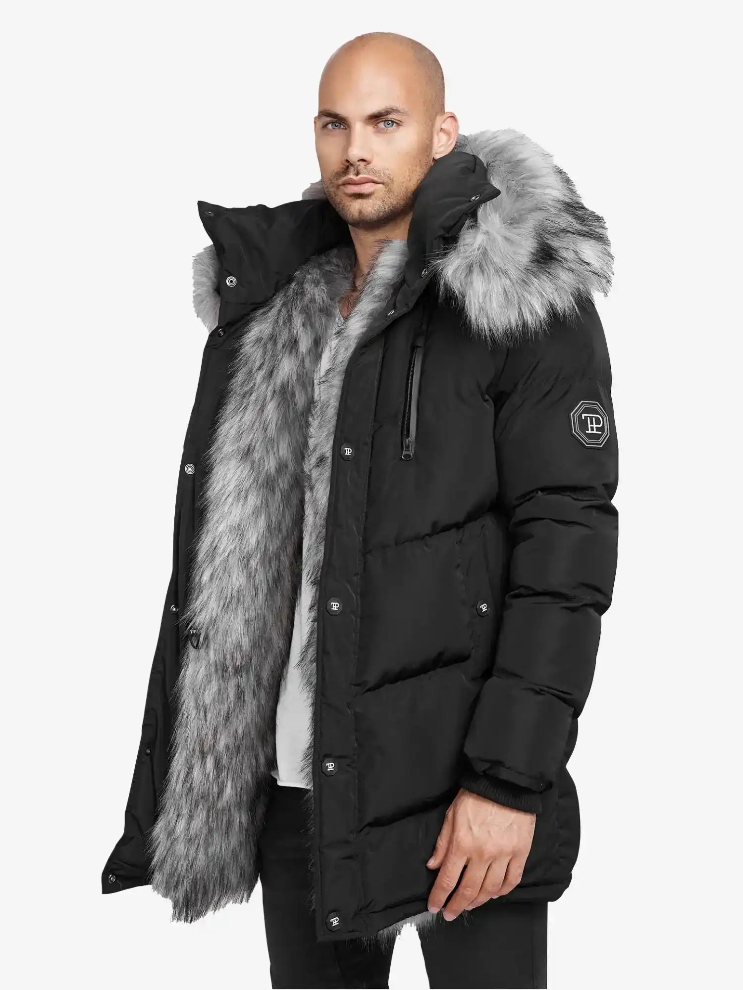 Ein Mann trägt die trueprodigy Zayn Winterjacke mit Kunstfell, einen schwarzen, wasserabweisenden Parka mit dickem grauem Kunstfell an Kapuze und Futter. Er ist mit einem Ärmelaufnäher und Druckknöpfen versehen und wird vor einem schlichten weißen Hintergrund gezeigt.