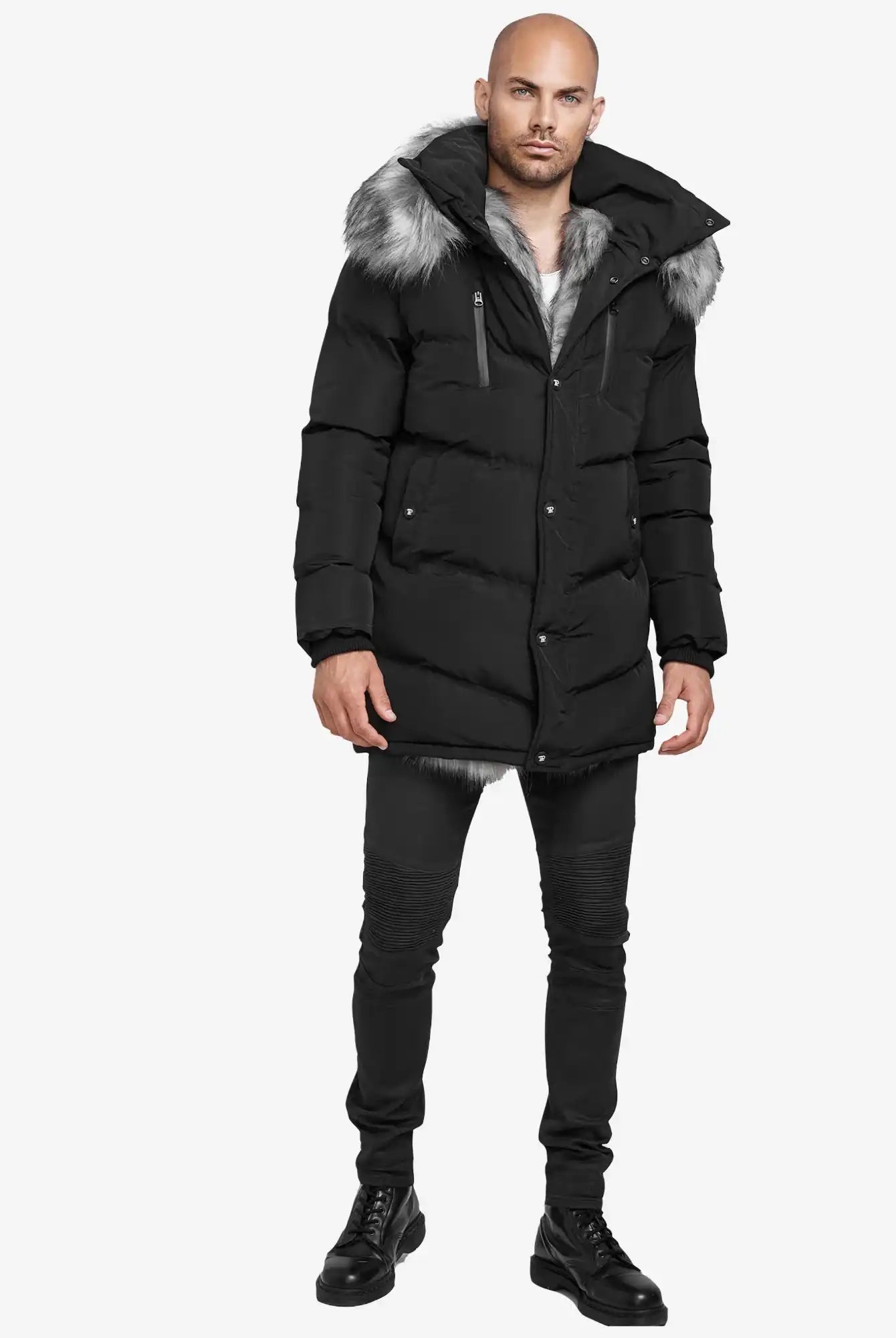 Ein Mann mit rasiertem Kopf und neutralem Gesichtsausdruck blickt vor einem schlichten weißen Hintergrund nach vorne. Er trägt die trueprodigy Zayn Winterjacke mit Kunstfell in Schwarz, gepaart mit einer schwarzen Jeans und Schnürstiefeln. Der Parka hat eine Kapuze mit Kunstfellbesatz.