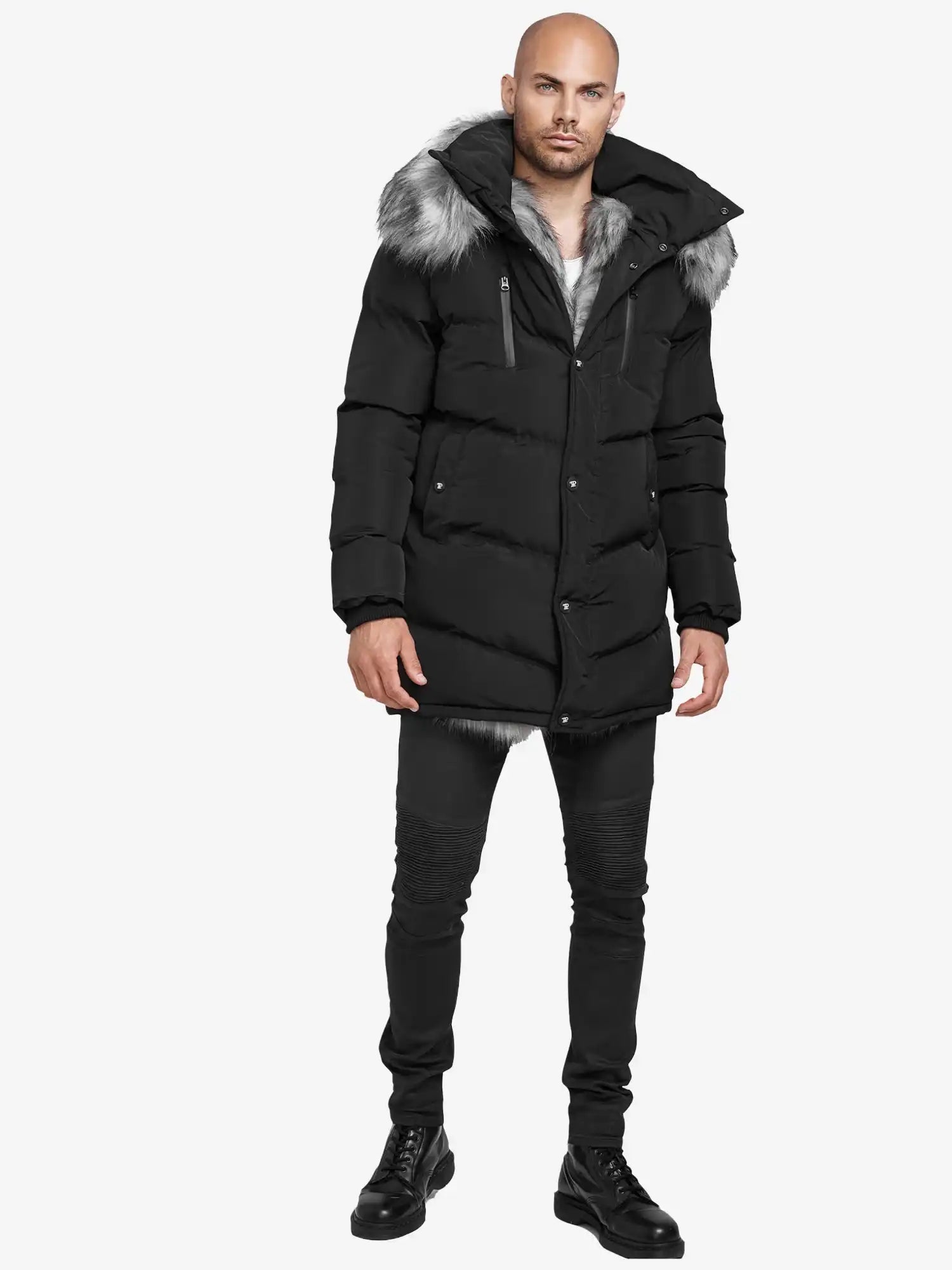Ein Mann mit rasiertem Kopf und neutralem Gesichtsausdruck blickt vor einem schlichten weißen Hintergrund nach vorne. Er trägt die trueprodigy Zayn Winterjacke mit Kunstfell in Schwarz, gepaart mit einer schwarzen Jeans und Schnürstiefeln. Der Parka hat eine Kapuze mit Kunstfellbesatz.