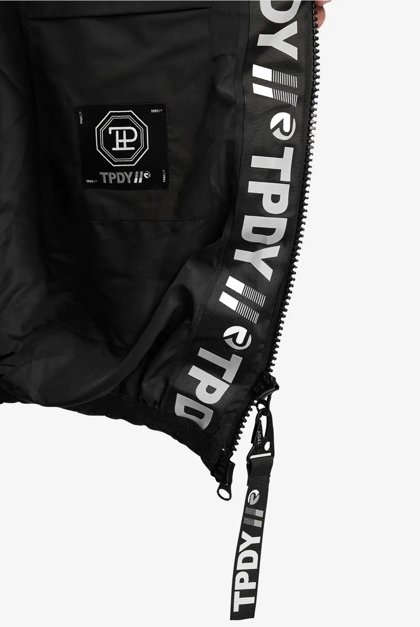 Nahaufnahme der Innenseite der Logan Pufferjacke mit Logo von trueprodigy. Hervorzuheben sind ein Reißverschluss, eine Tasche mit sechseckigem Logoaufnäher und wiederholter weißer TPDY-Text und -Symbole entlang der Innenverkleidung und des Reißverschlusses.