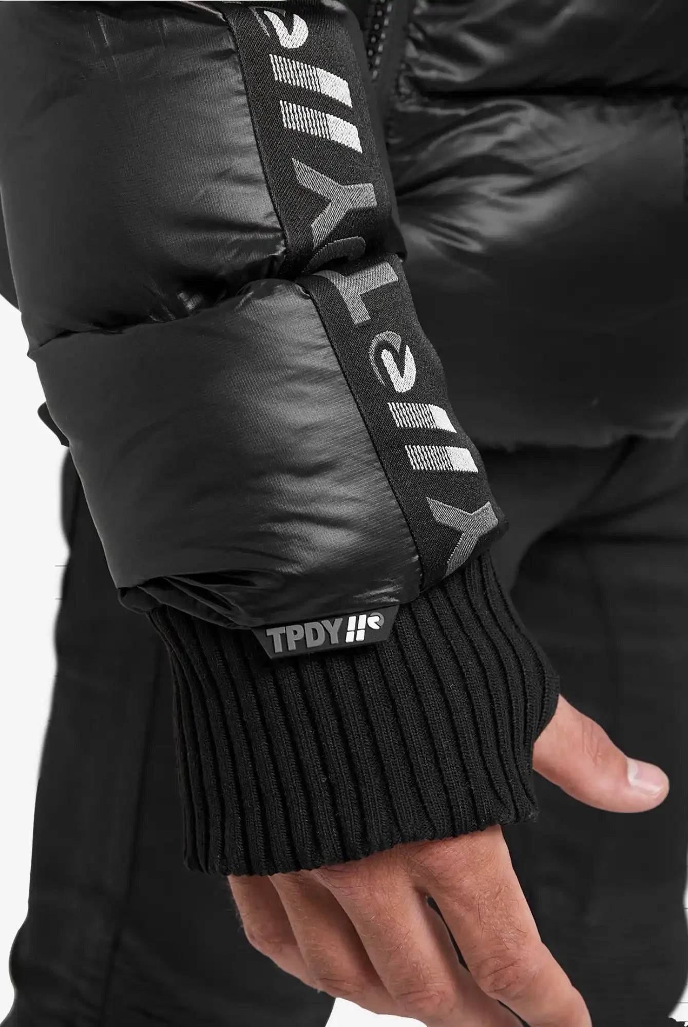 Eine Nahaufnahme einer Hand, die die trueprodigy Logan Pufferjacke mit Logo trägt, zeigt Rippstrickbündchen und einen aufgedruckten weißen Text, darunter TPDY und stilisierte Symbole, auf dem Ärmel.