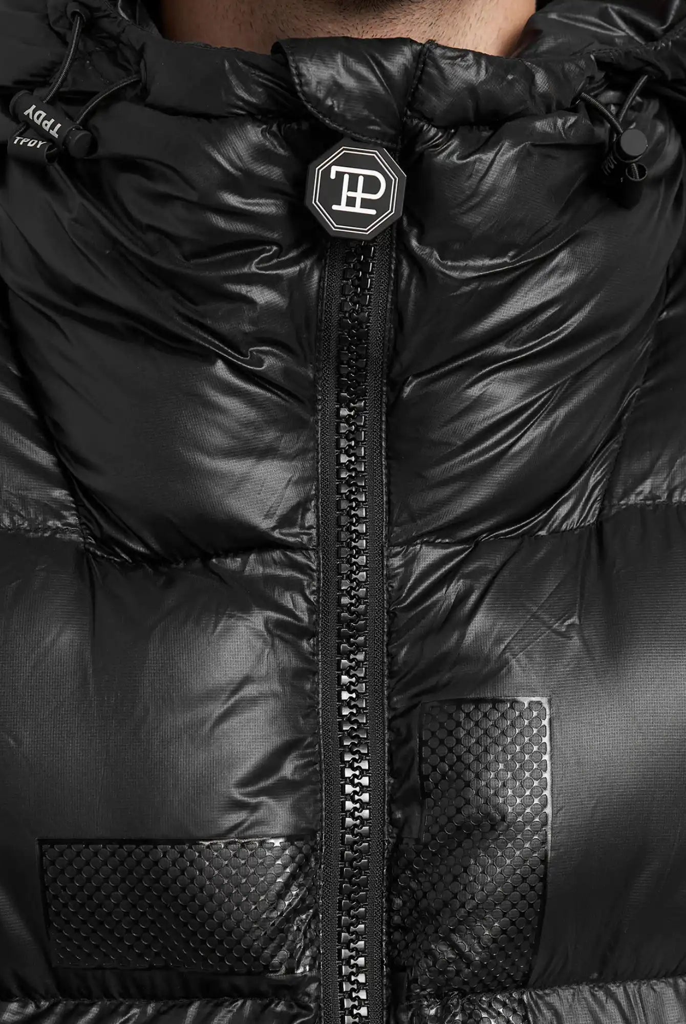 Nahaufnahme der trueprodigy Logan Pufferjacke mit Logo, mit zentralem Reißverschluss, strukturierten quadratischen Patches, einem runden Logo-Badge am Kragen, glänzendem Finish und sichtbaren Nähten.