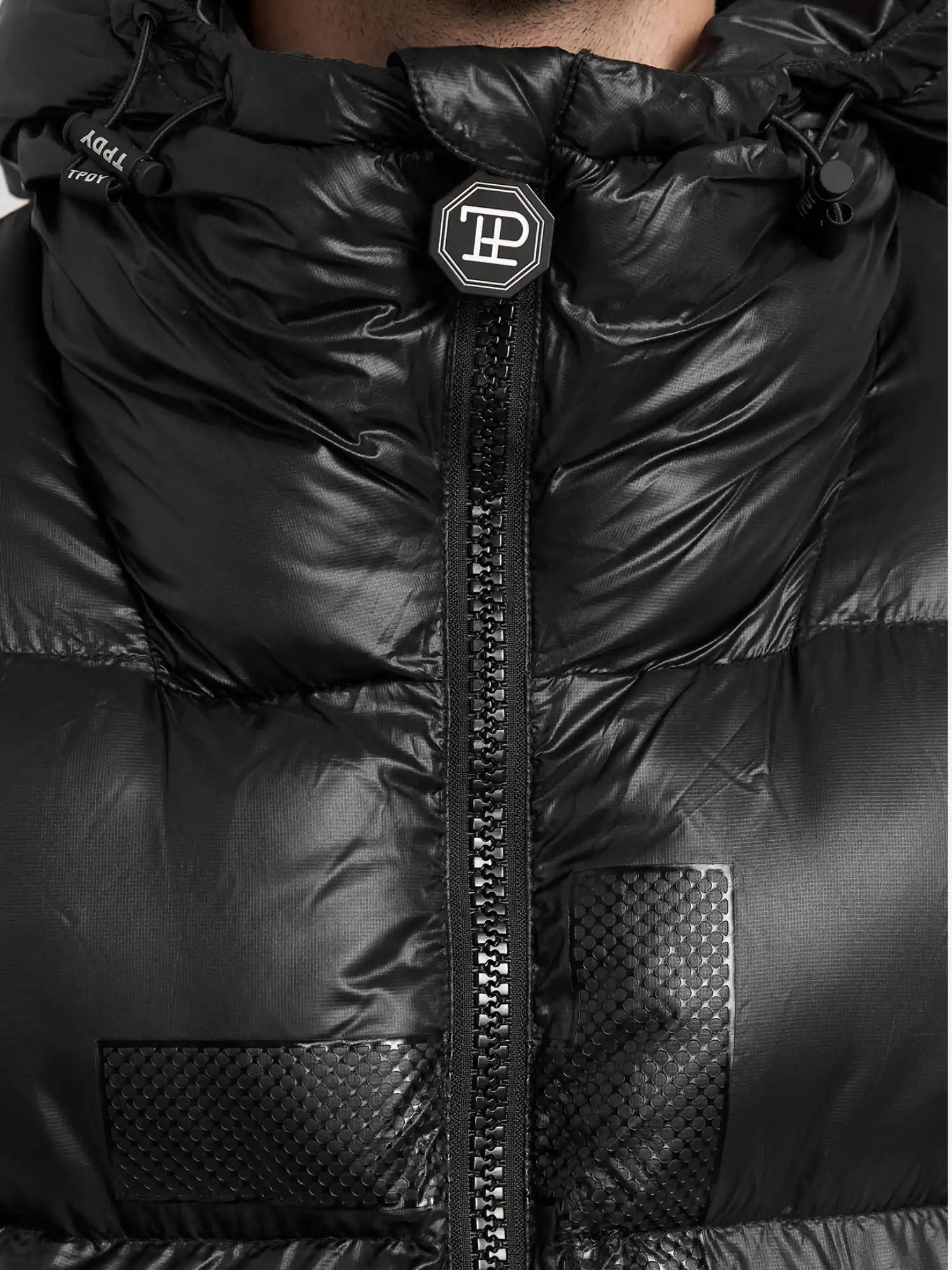 Nahaufnahme der trueprodigy Logan Pufferjacke mit Logo, mit zentralem Reißverschluss, strukturierten quadratischen Patches, einem runden Logo-Badge am Kragen, glänzendem Finish und sichtbaren Nähten.