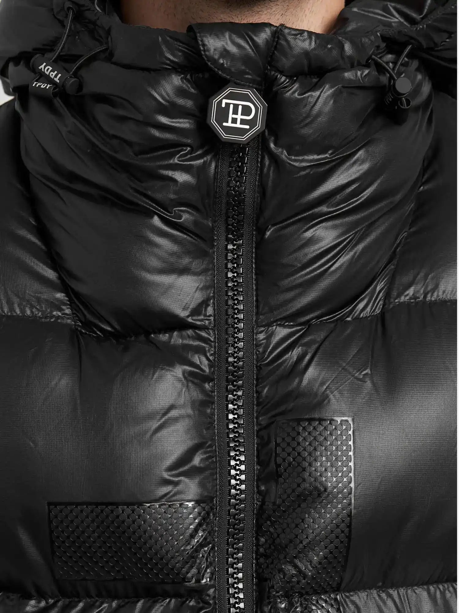 Nahaufnahme der trueprodigy Logan Pufferjacke mit Logo, mit zentralem Reißverschluss, strukturierten quadratischen Patches, einem runden Logo-Badge am Kragen, glänzendem Finish und sichtbaren Nähten.