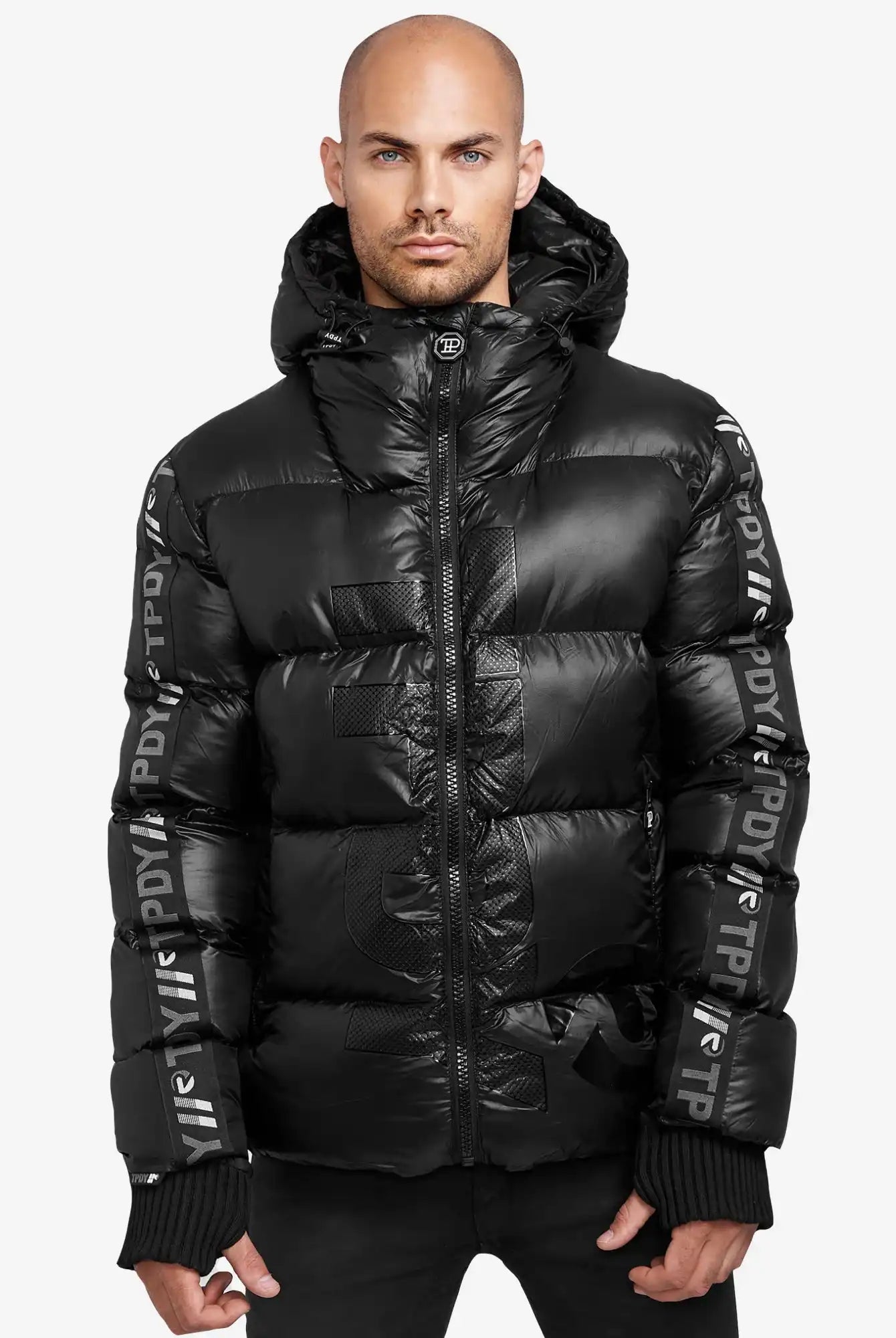 Ein Mann mit rasiertem Kopf trägt die trueprodigy Logan Pufferjacke mit Logo in glänzendem Schwarz, mit sichtbaren Logos und Ärmeltext. Er steht mit neutralem Gesichtsausdruck vor einem schlichten weißen Hintergrund.