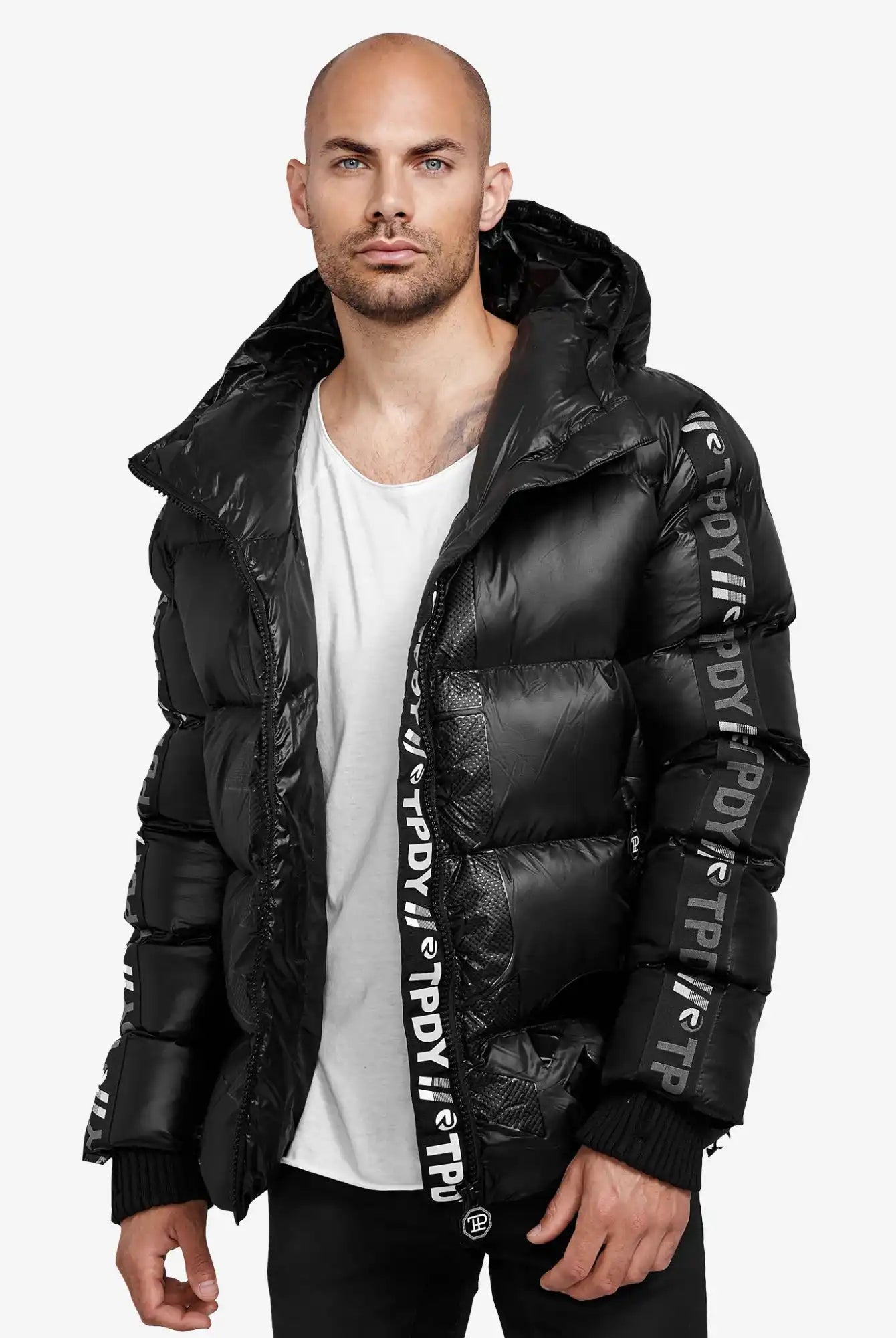 Ein Mann mit gestutztem Bart und rasiertem Kopf trägt die trueprodigy Logan Pufferjacke mit Logo, gepaart mit einem weißen Hemd und einer schwarzen Hose, vor einem schlichten Hintergrund.