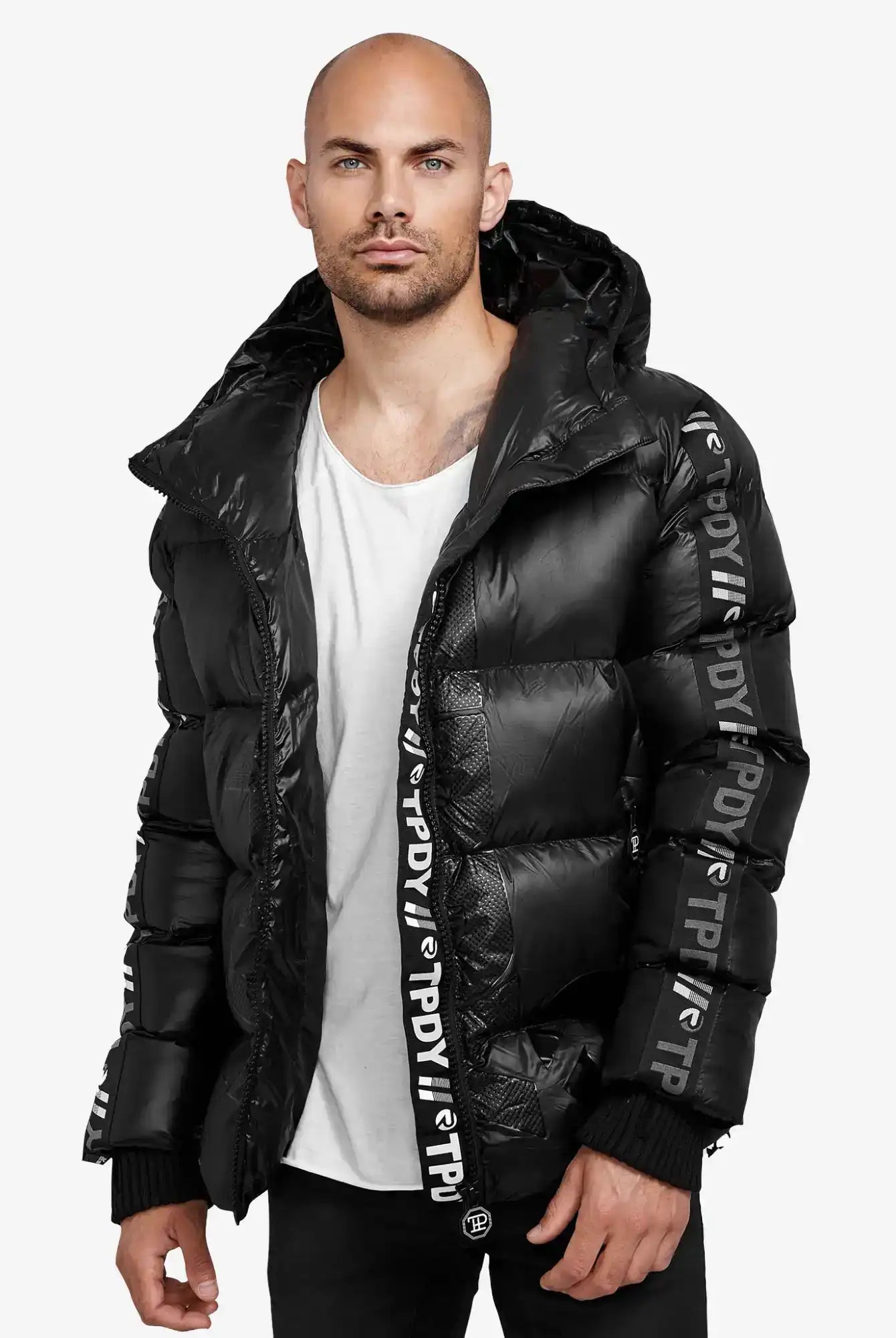 Ein Mann mit gestutztem Bart und rasiertem Kopf trägt die trueprodigy Logan Pufferjacke mit Logo, gepaart mit einem weißen Hemd und einer schwarzen Hose, vor einem schlichten Hintergrund.