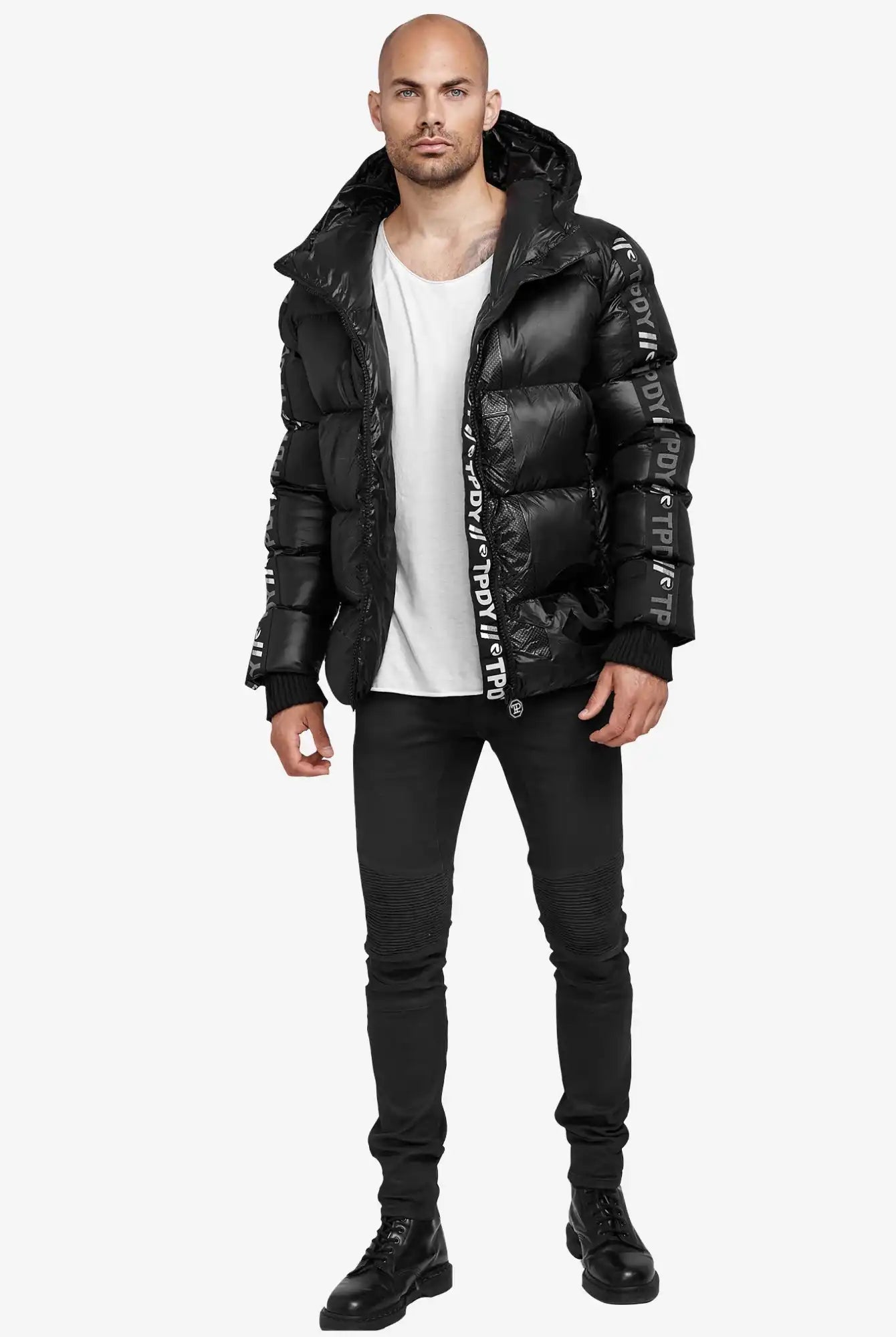 Ein Mann blickt nach vorne in einer teilweise geöffneten Logan Pufferjacke mit Logo von trueprodigy mit Textdetails über einem weißen T-Shirt, schwarzen Skinny Jeans und schwarzen Stiefeln vor einem schlichten weißen Hintergrund.